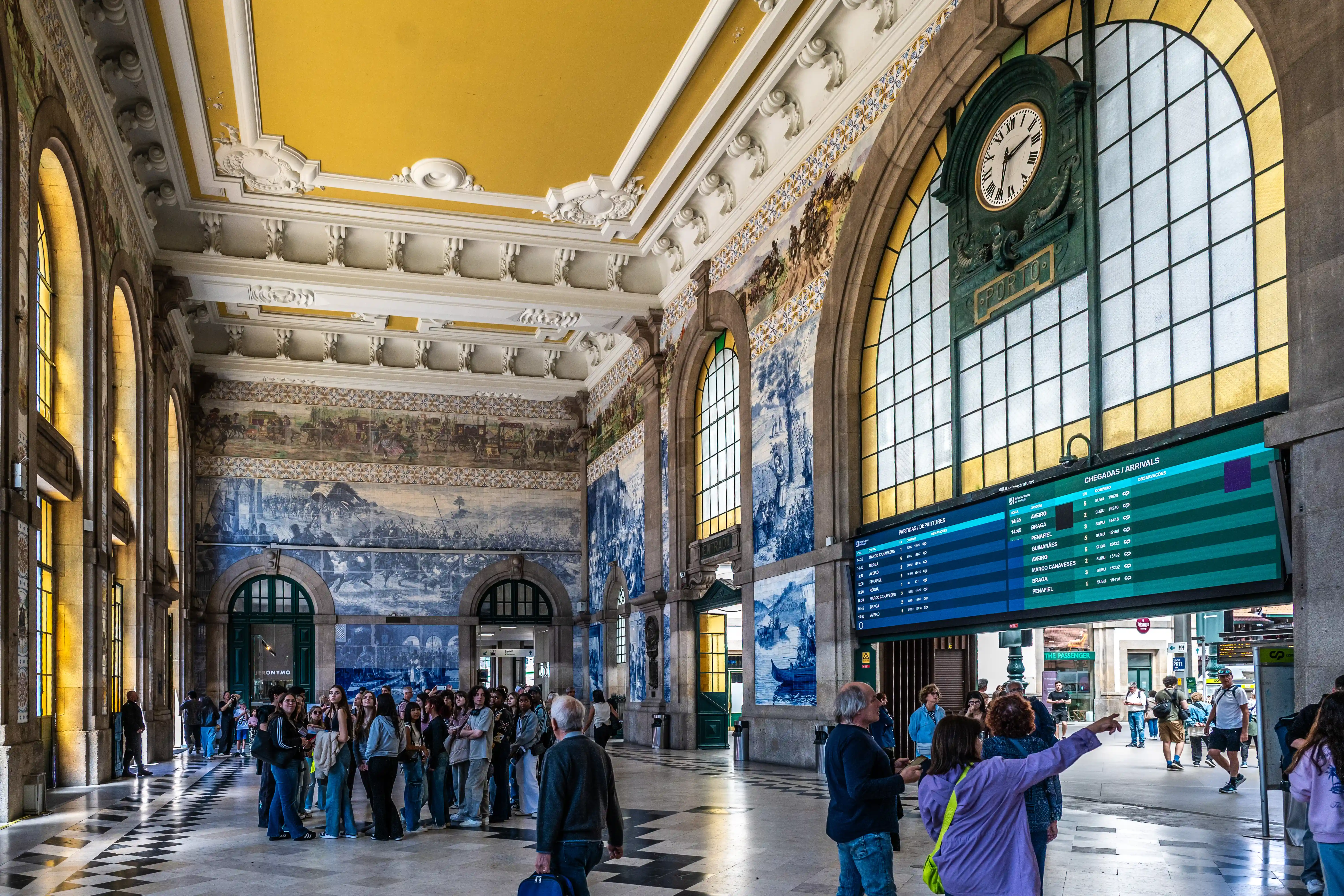 A bustling interior of a grand train station hall adorned with intricate blue and white azulejo tiles, featuring a large digital departures board, a classic clock, and sunlight streaming through arched windows.