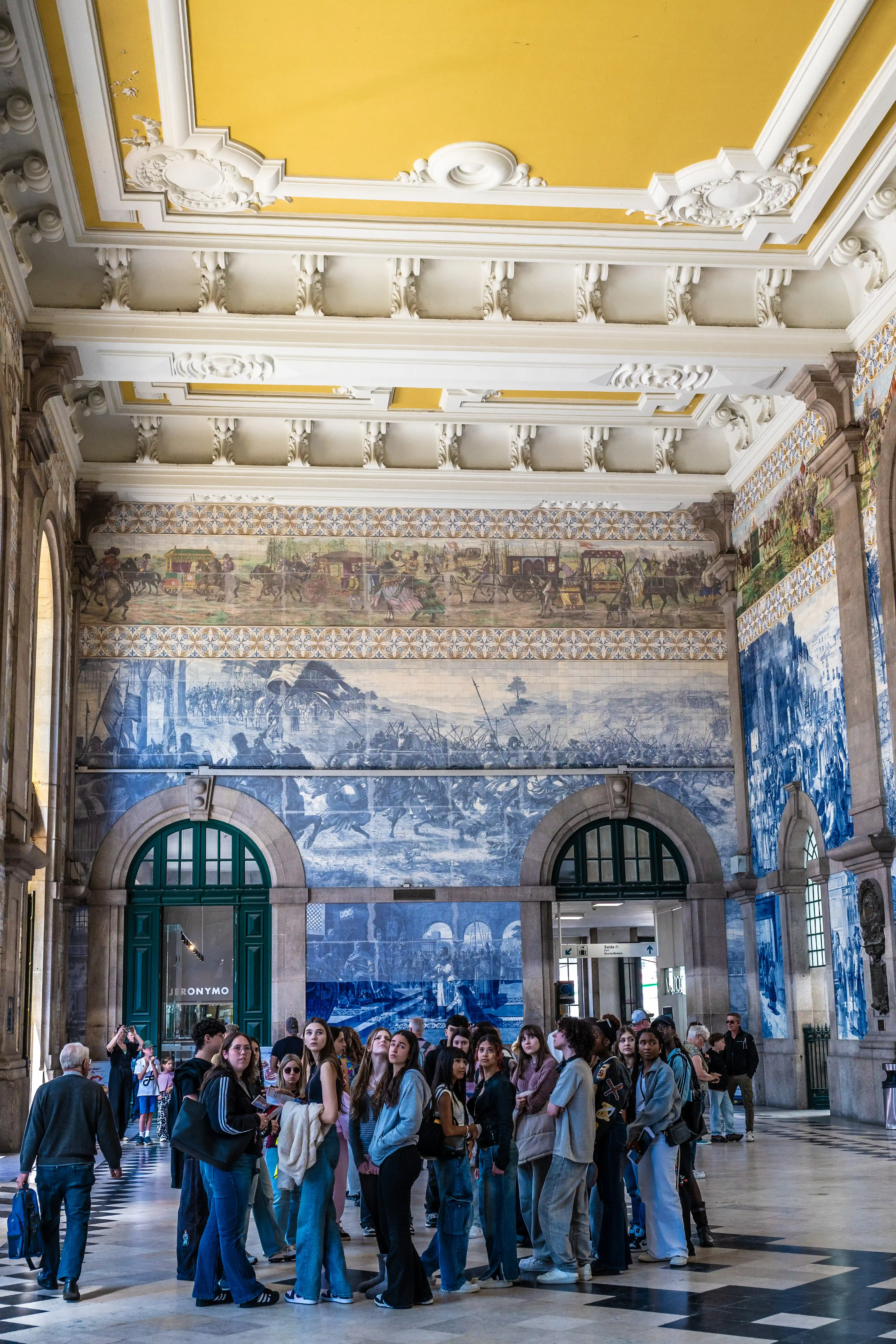 A crowd of people, predominantly young adults, stand in a large, well-lit hall of a train station. The walls are covered in large blue and white tiled murals that depict historical battle scenes and processions. Above the murals, a decorative frieze runs along the wall, topped by a richly ornamented ceiling painted in yellow and white. Several arched doorways and windows are visible, some with dark green doors. The floor is a patterned marble.