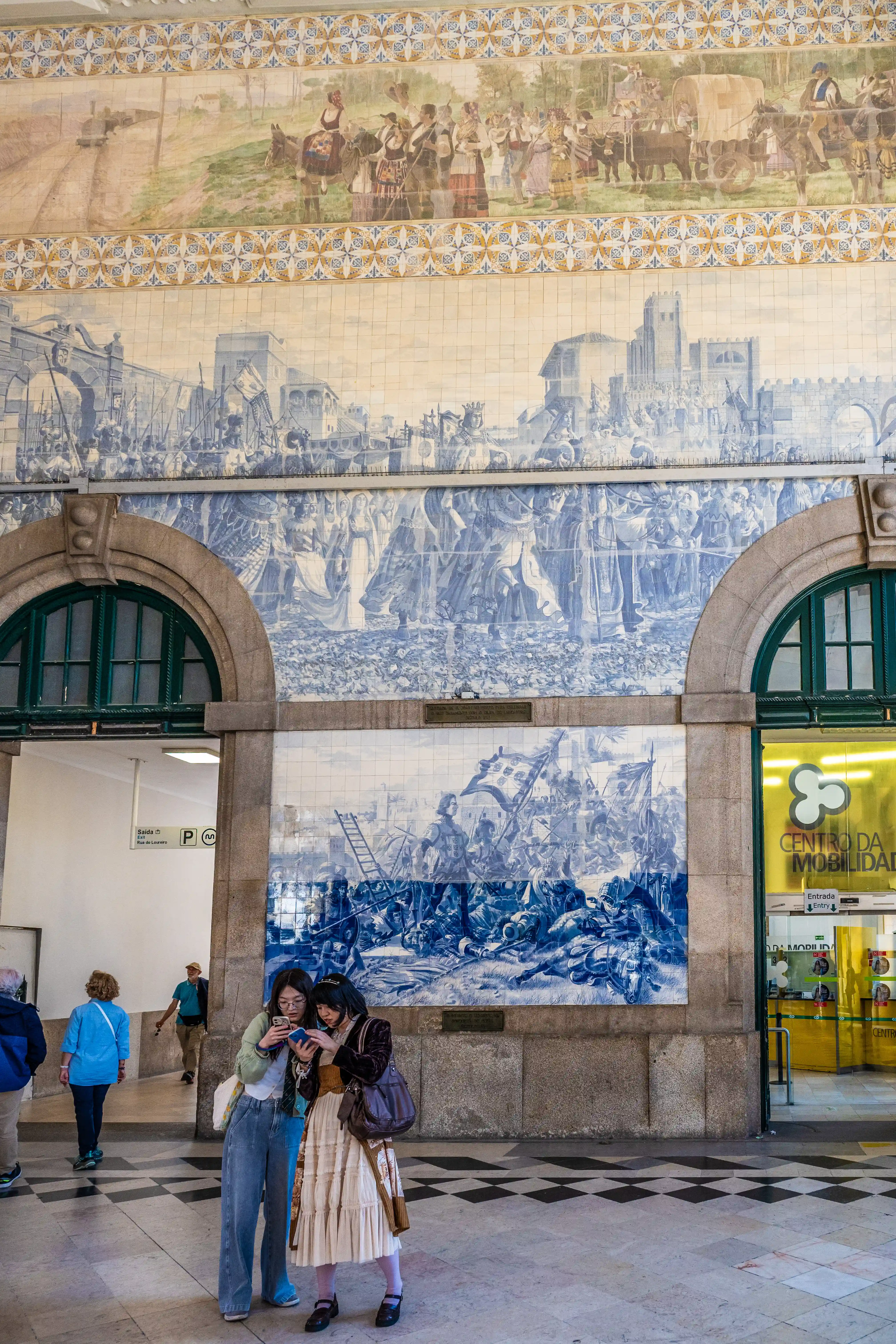 Two young women look at a mobile phone in front of a vast, multi-panelled azulejo mural in a train station. The blue and white tiles depict detailed historical scenes, including a procession at the top and battle scenes below. Arched doorways and a modern Centro da Mobilidade entrance are visible.
