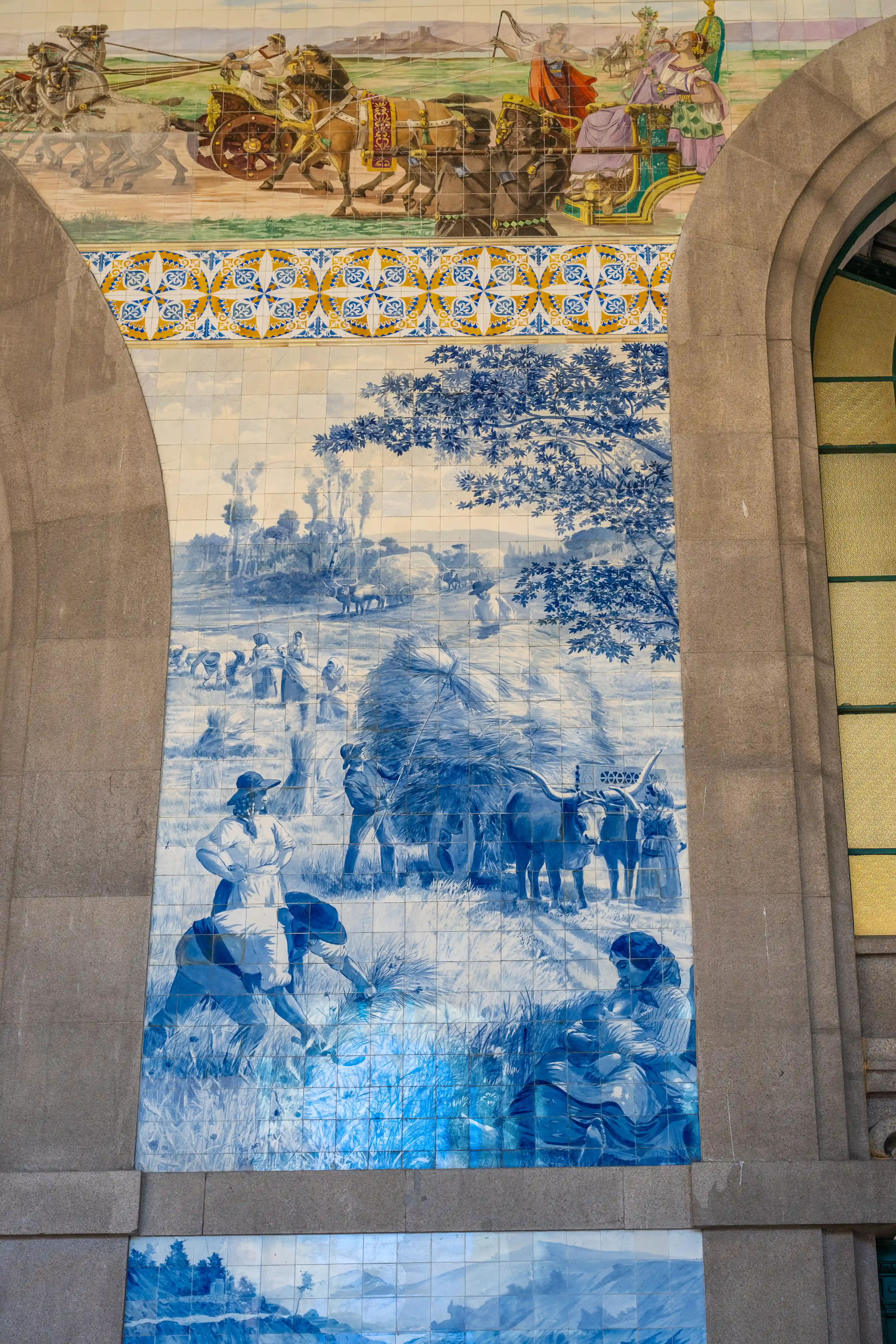 A grand tiled mural in blue and white depicting a busy rural harvest scene with people, oxen, and carts, set beneath a colourful frieze of Roman chariots pulled by horses.