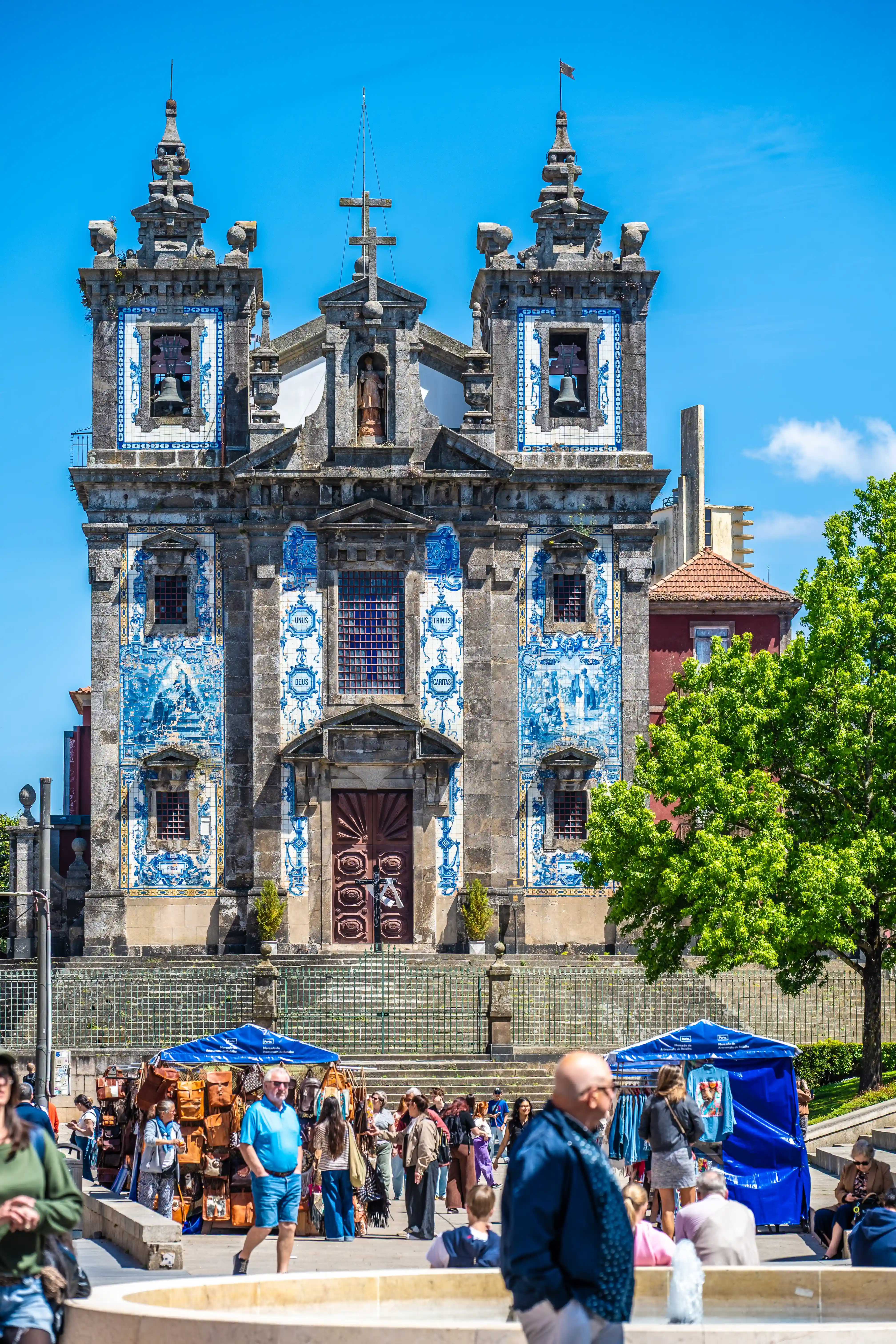 A detailed shot of the Igreja Paroquial de Santo Ildefonso, Portugal, featuring its elaborate azulejo-tiled facade and twin bell towers. In front of the church, a busy outdoor market is in full swing with stalls selling bags and clothing, and people are seen shopping and walking around.