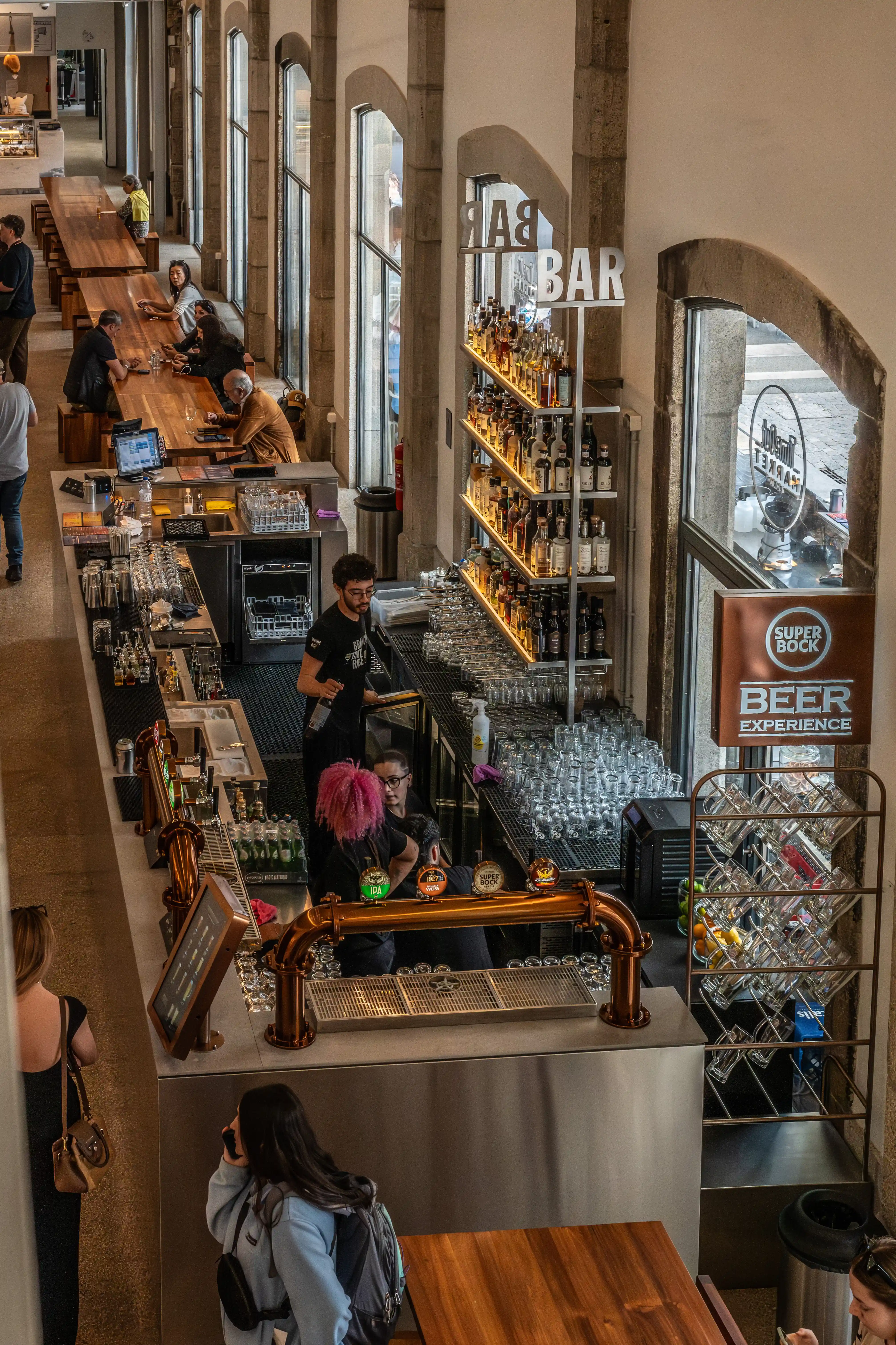 Overhead view of a busy bar and dining area with people sitting at long wooden tables, a bar stocked with drinks, and beer taps.