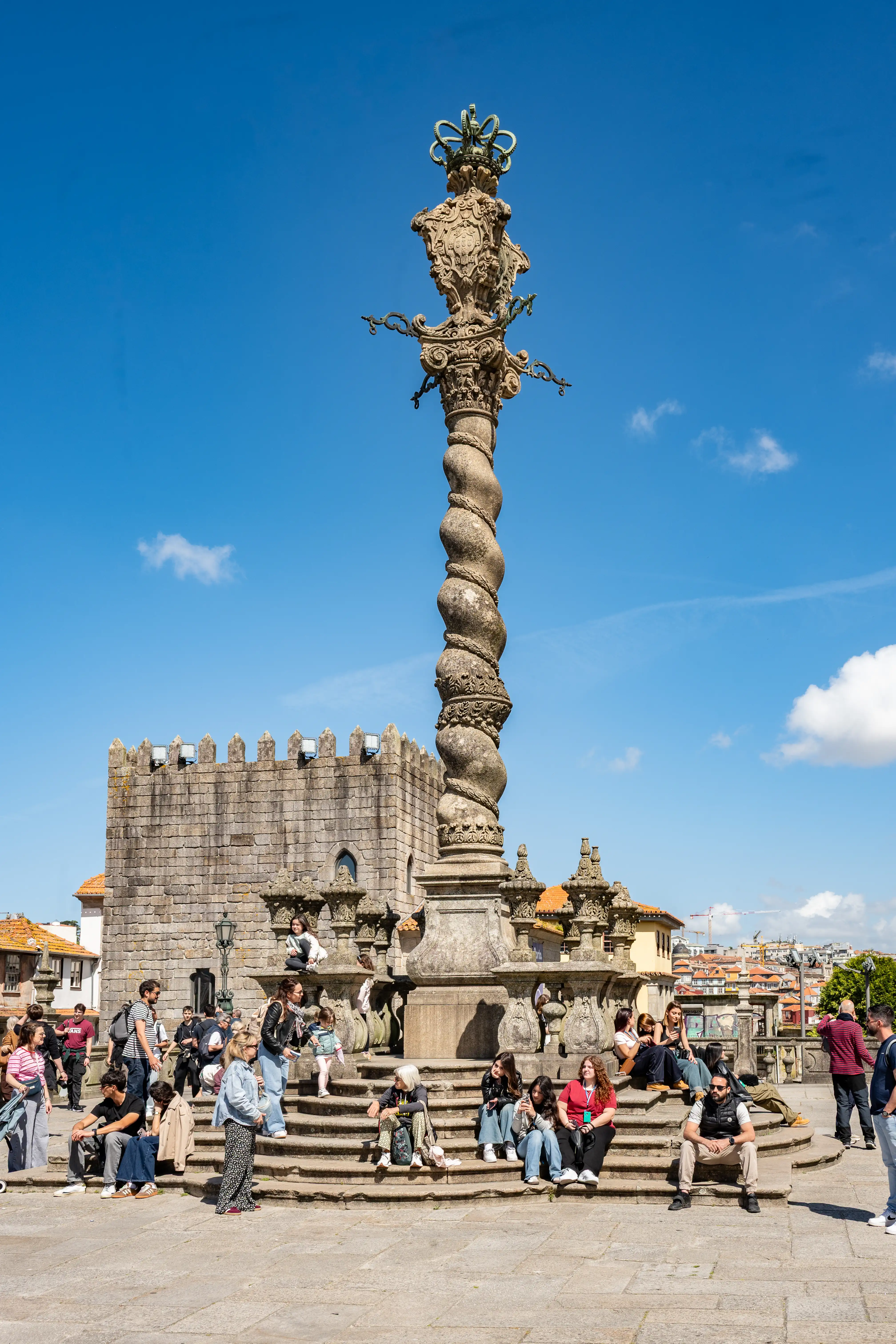 A spiral stone pillory topped with a crown stands in a square in Porto, with people sitting on the steps and a historic stone building in the background.