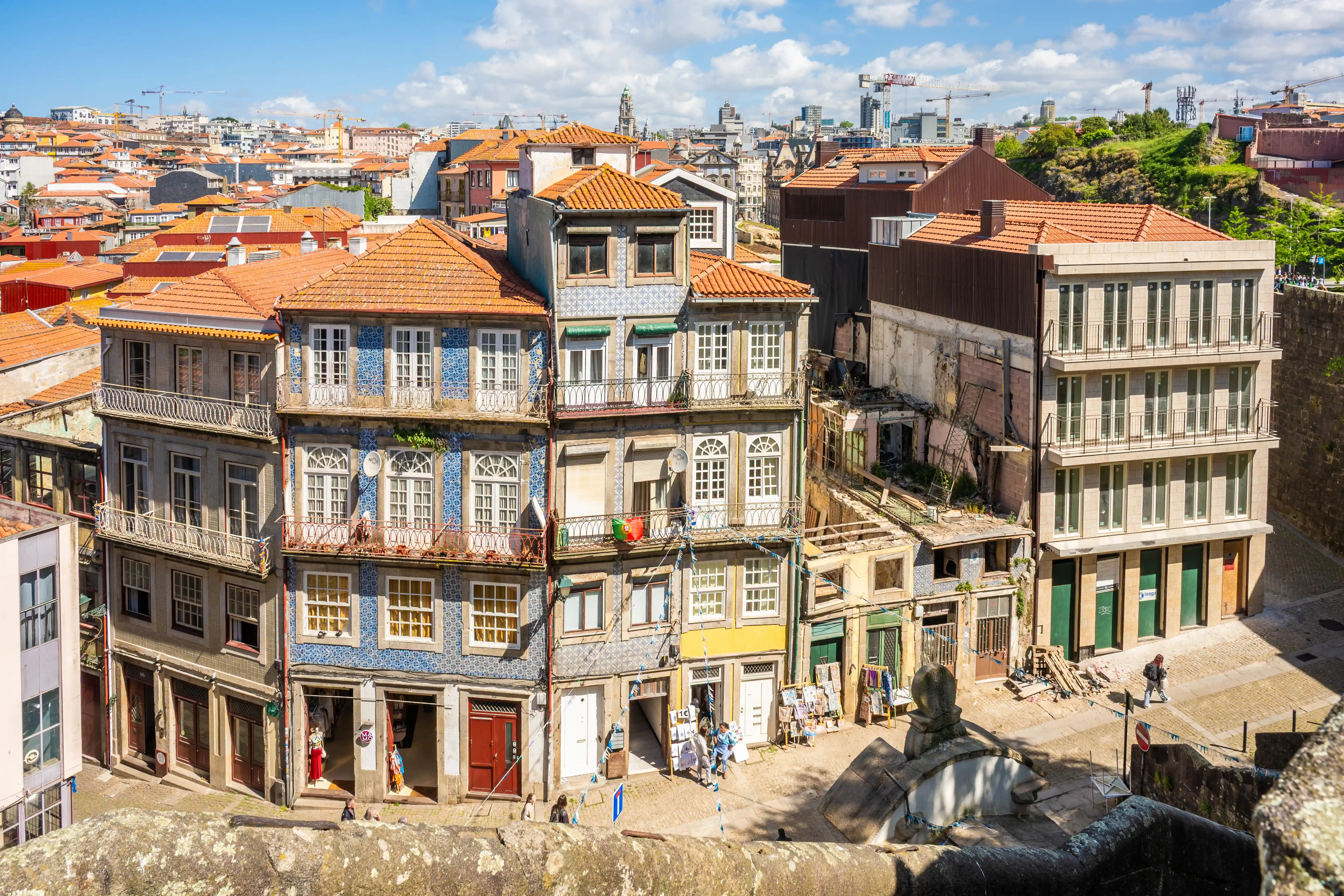 High-angle view of closely packed buildings with red-tiled roofs in a European city, likely Porto, under a partly cloudy sky, with a stone wall in the foreground.