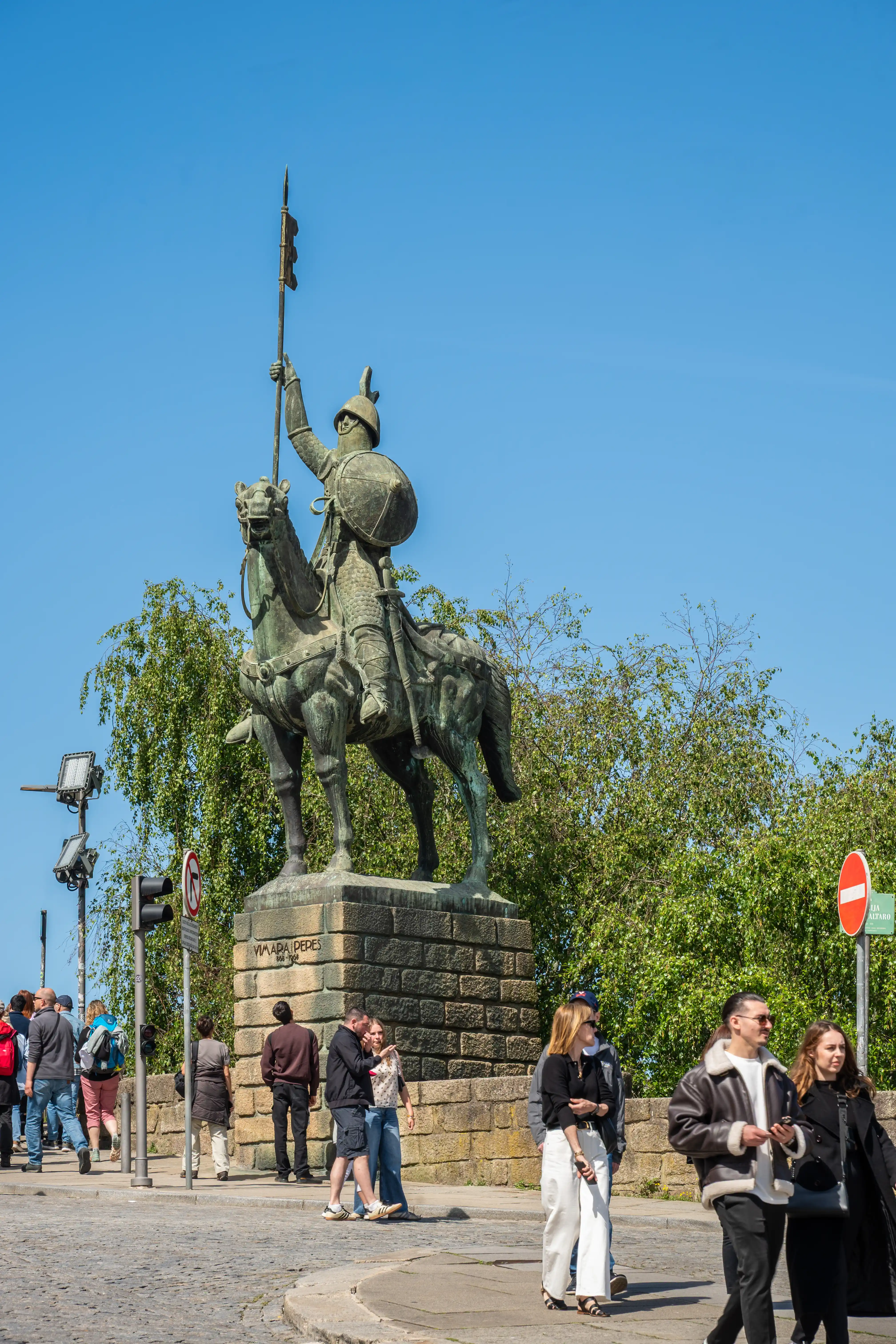 Bronze statue of a knight on horseback, with people walking on a bridge in the foreground and trees and blue sky in the background.
