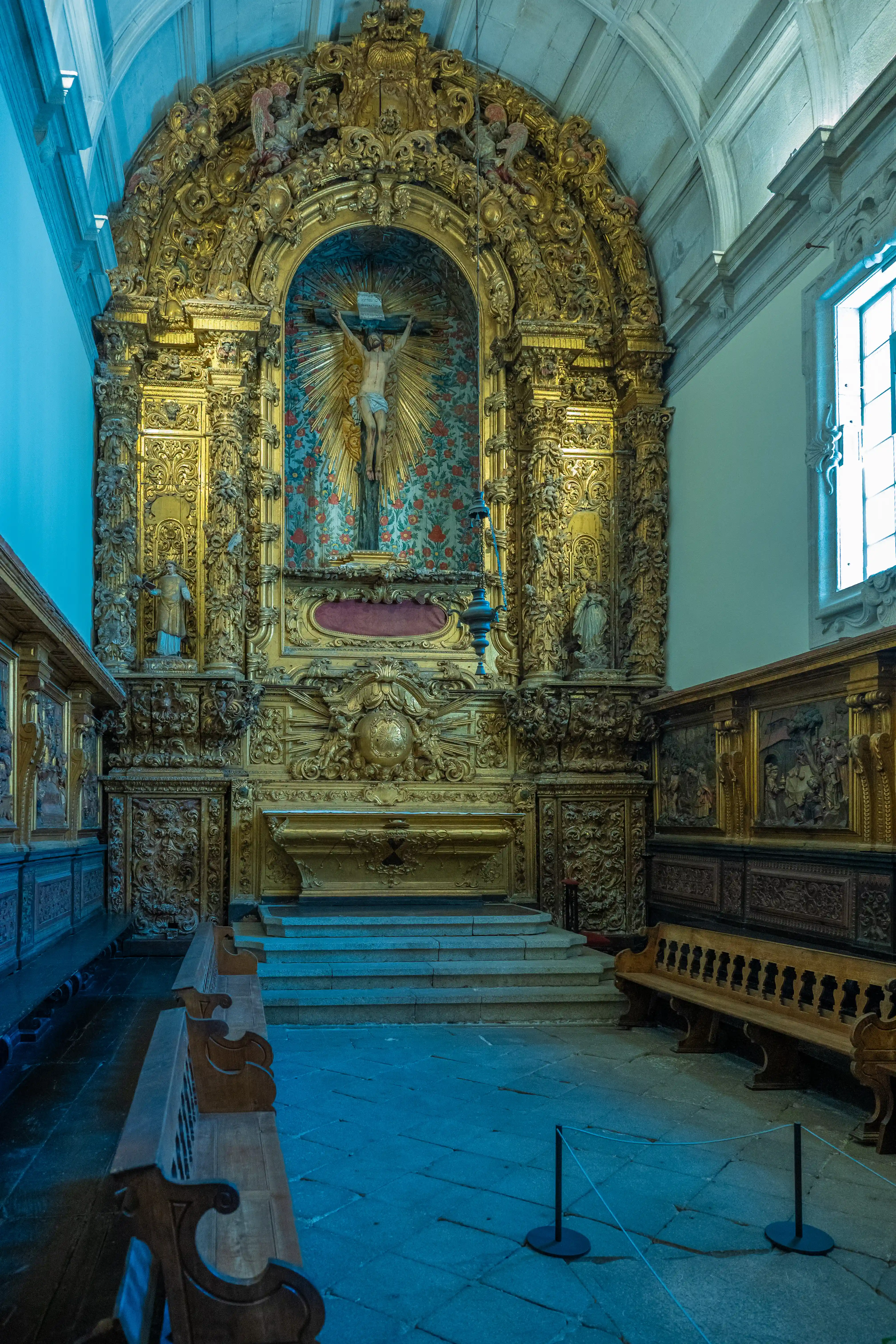 Interior view of a church chapel with a highly ornate, gilded Baroque altar. A statue of the crucified Christ is at the centre, surrounded by elaborate carvings and decorative elements. Wooden pews are in the foreground, and a large window is visible on the right.