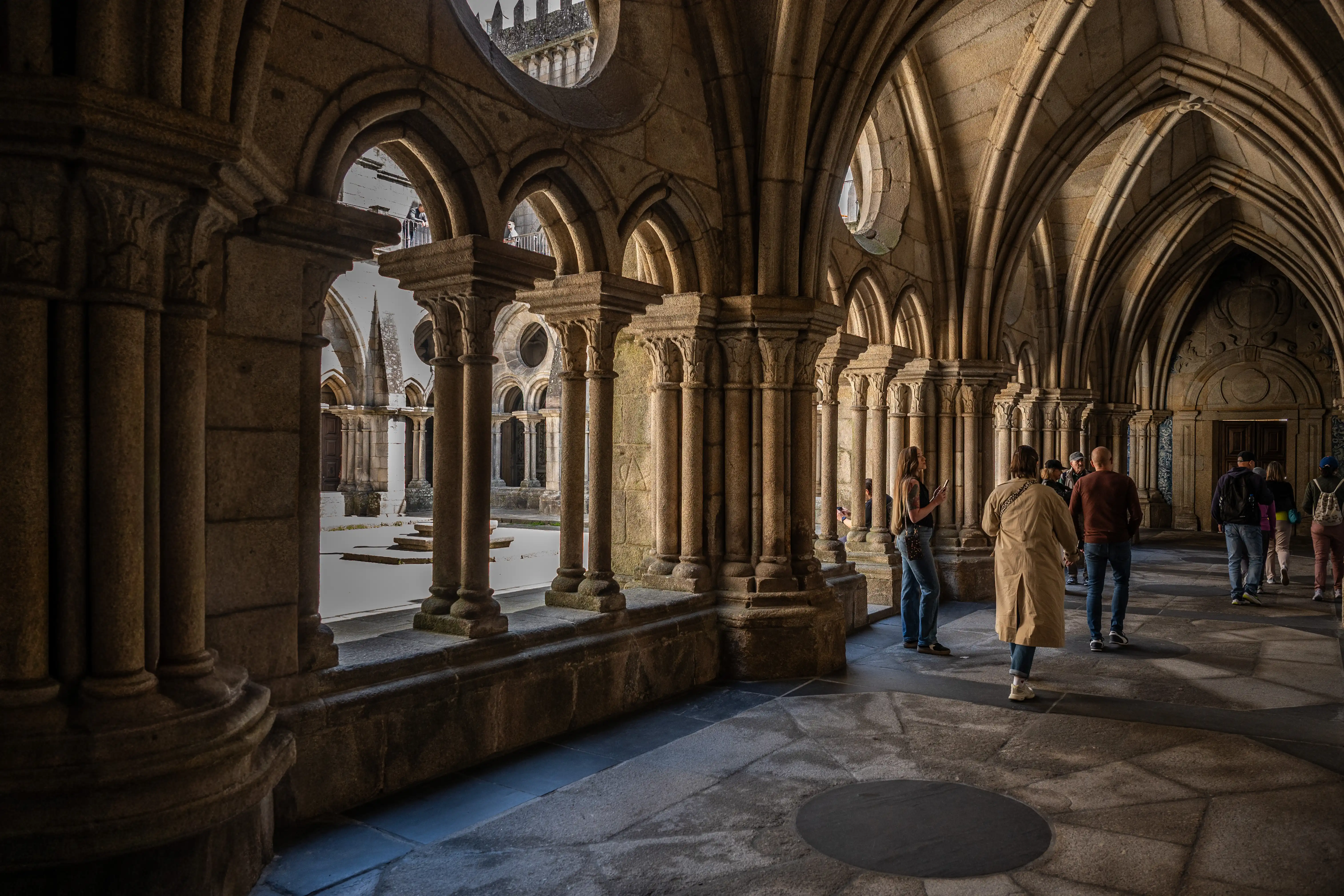 Silhouettes of tourists walking through a sunlit stone cloister with arched walkways and ornate columns.