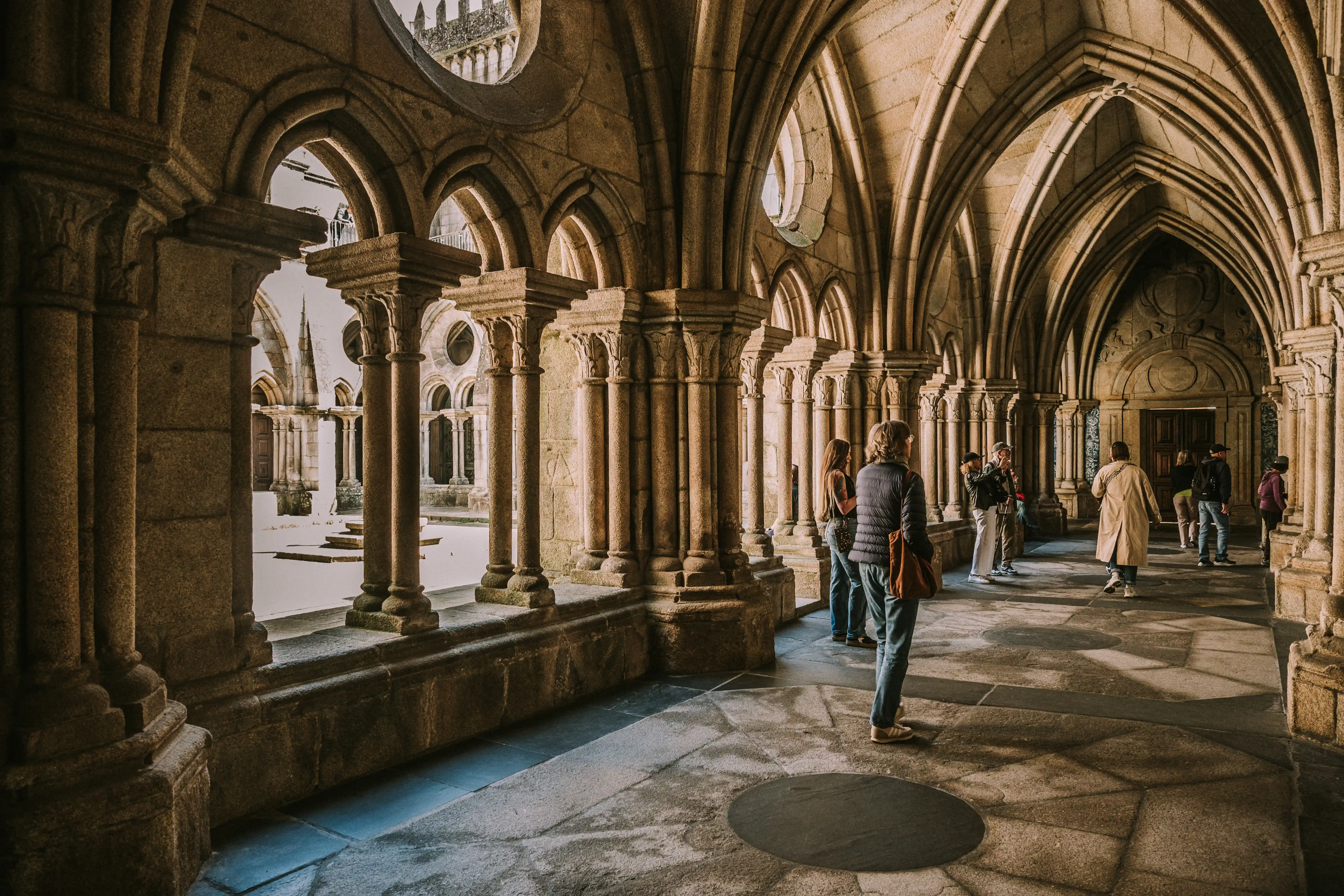 Tourists wander through a historic stone cloister with arched walkways and sunlight filtering in, highlighting the detailed architecture.