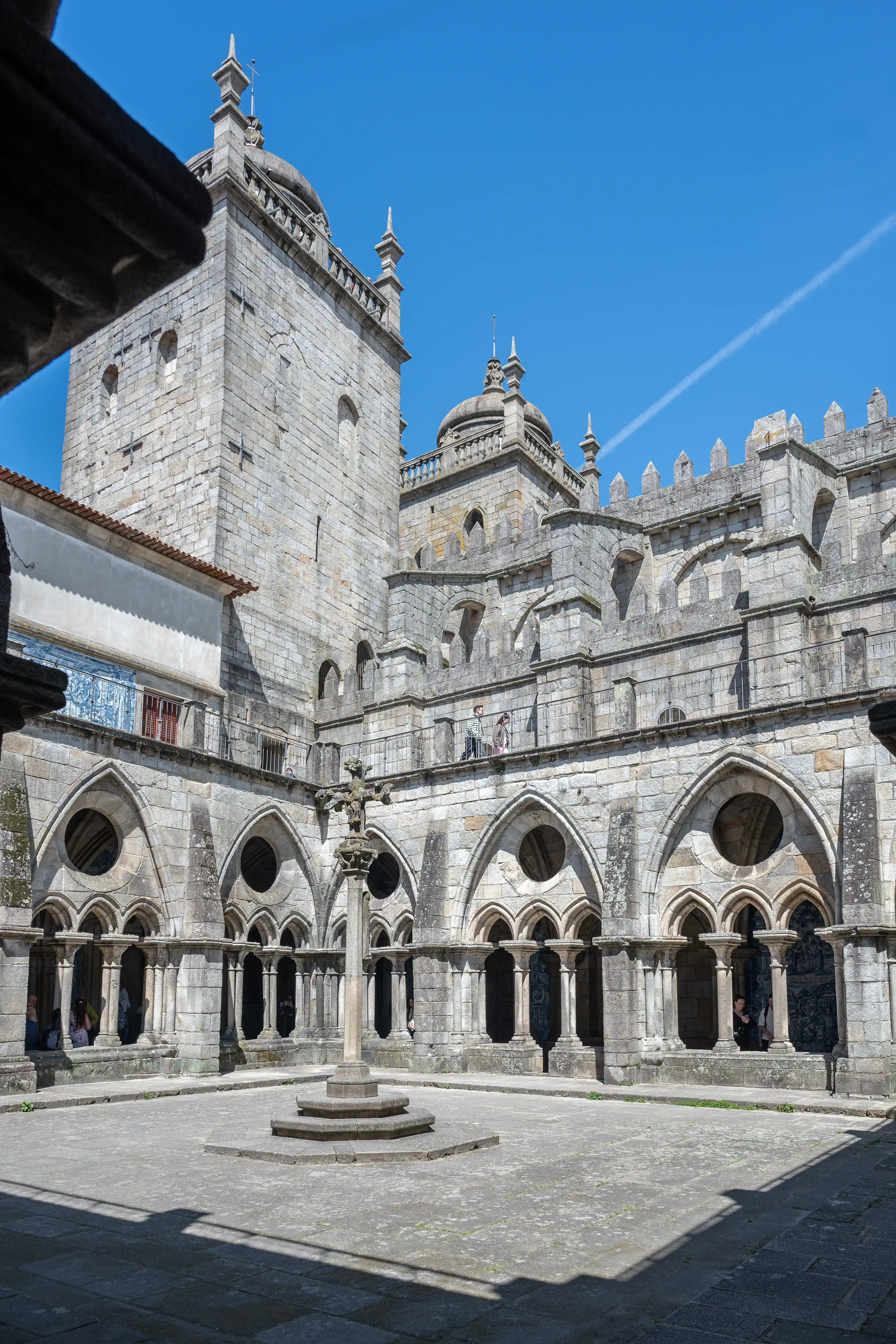 A sunlit courtyard of an ancient stone cathedral, featuring arched cloisters, a central stone cross, and towering cathedral spires against a blue sky.