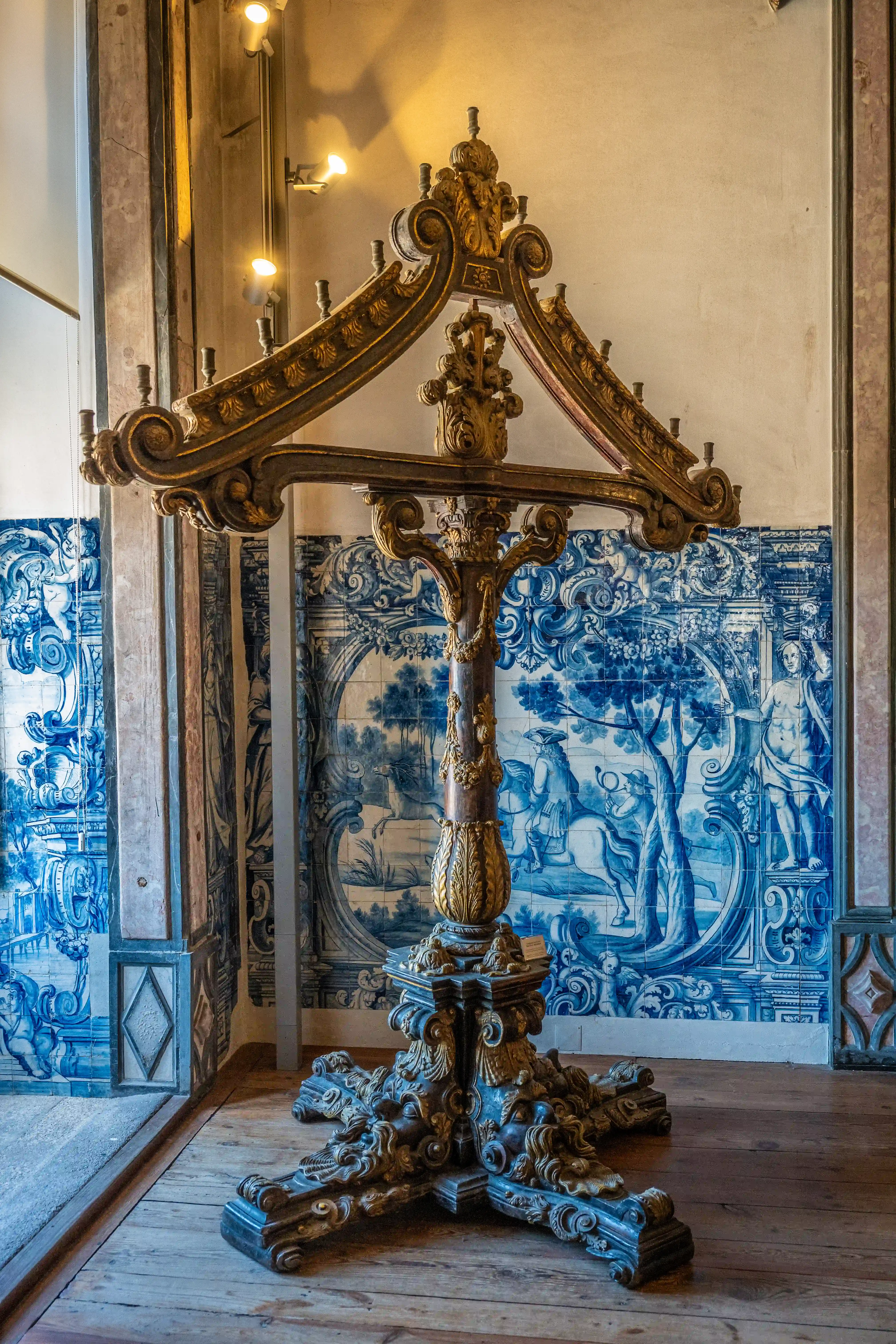 An ornate, gilded candelabra with a decorative pedestal stands on a wooden floor in front of a wall covered in blue and white Portuguese azulejo tiles. The tiles feature a detailed scenic depiction.