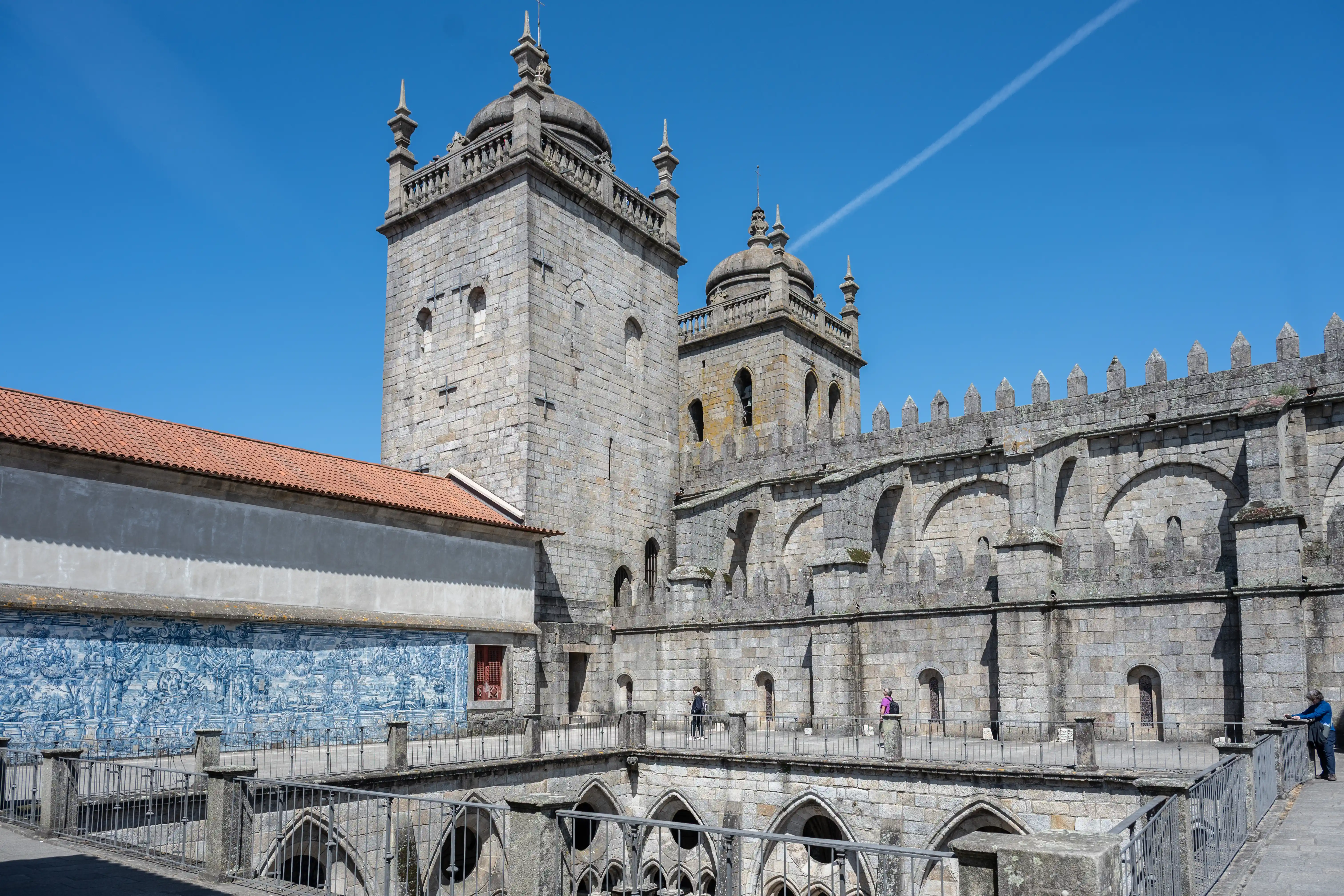 A wide shot of the Porto Cathedral with its stone towers, arched windows, and a tiled facade under a clear blue sky.