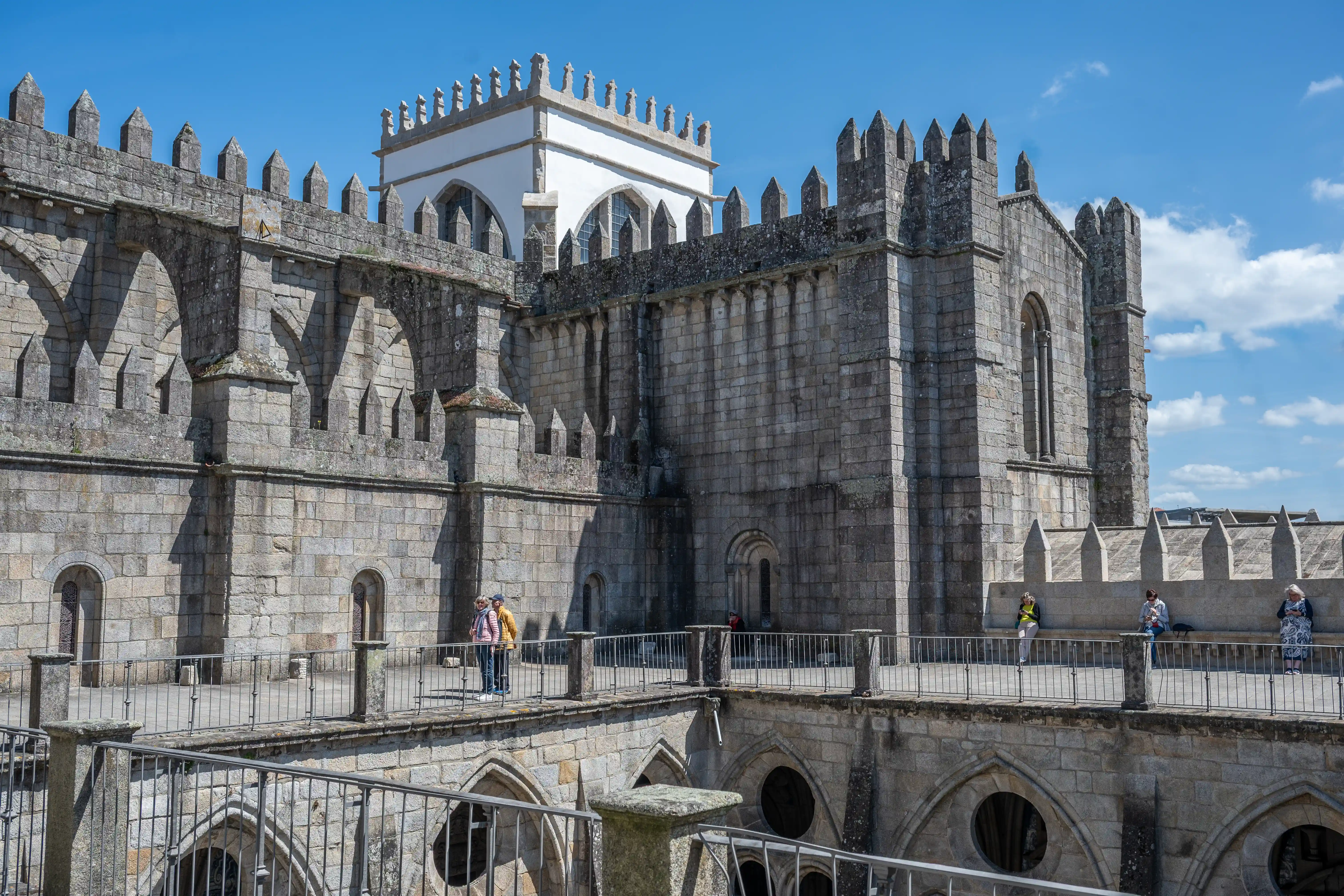  A large, grey stone cathedral with a white turret structure. Crenellated walls and arched walkways are prominent. Several people are walking on the walkways. A construction crane is in the sky in the background.
