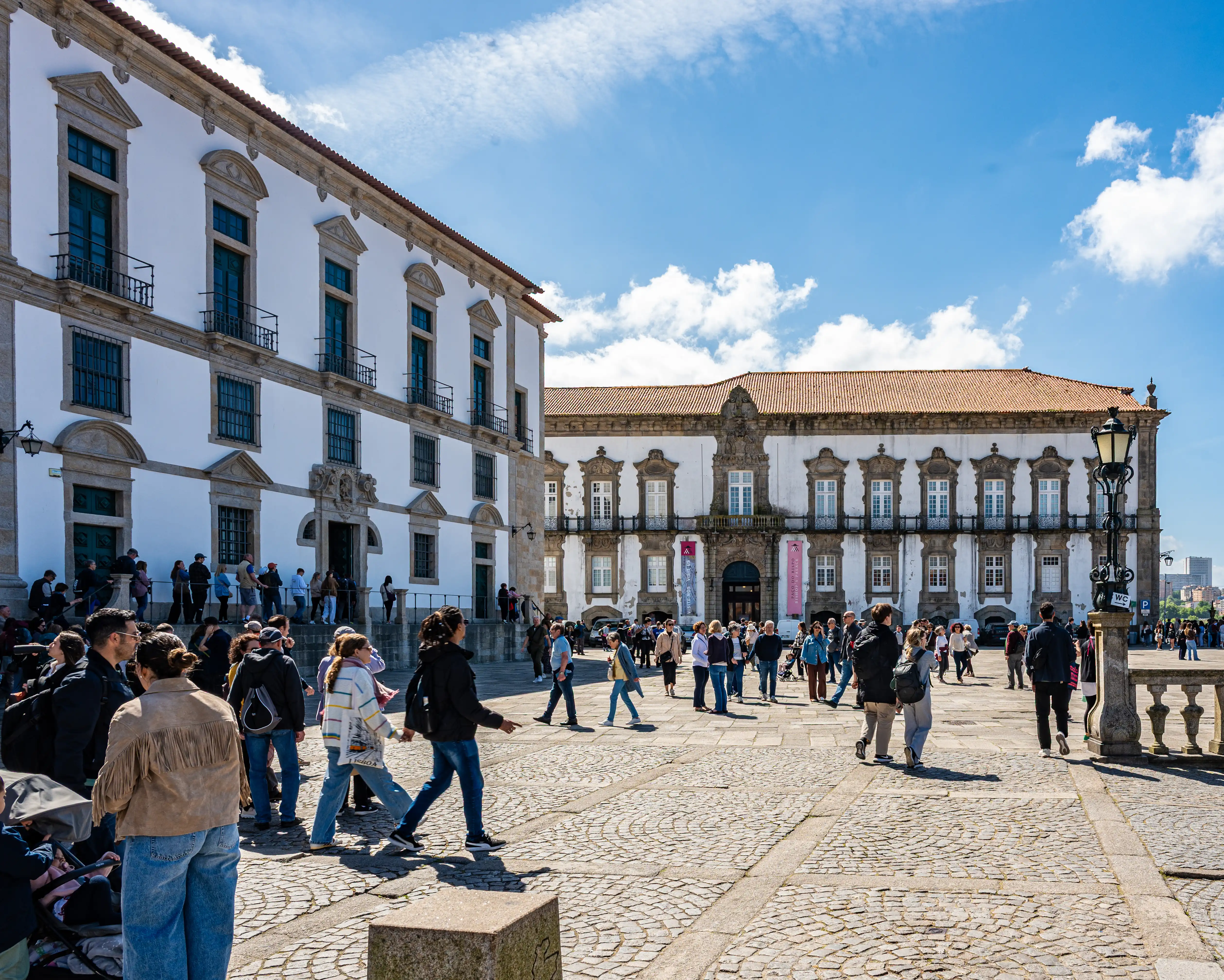 A crowded, cobblestone plaza in front of large, classical white buildings with terracotta roofs, under a bright blue sky with some clouds. People are seen walking and gathering in the square.