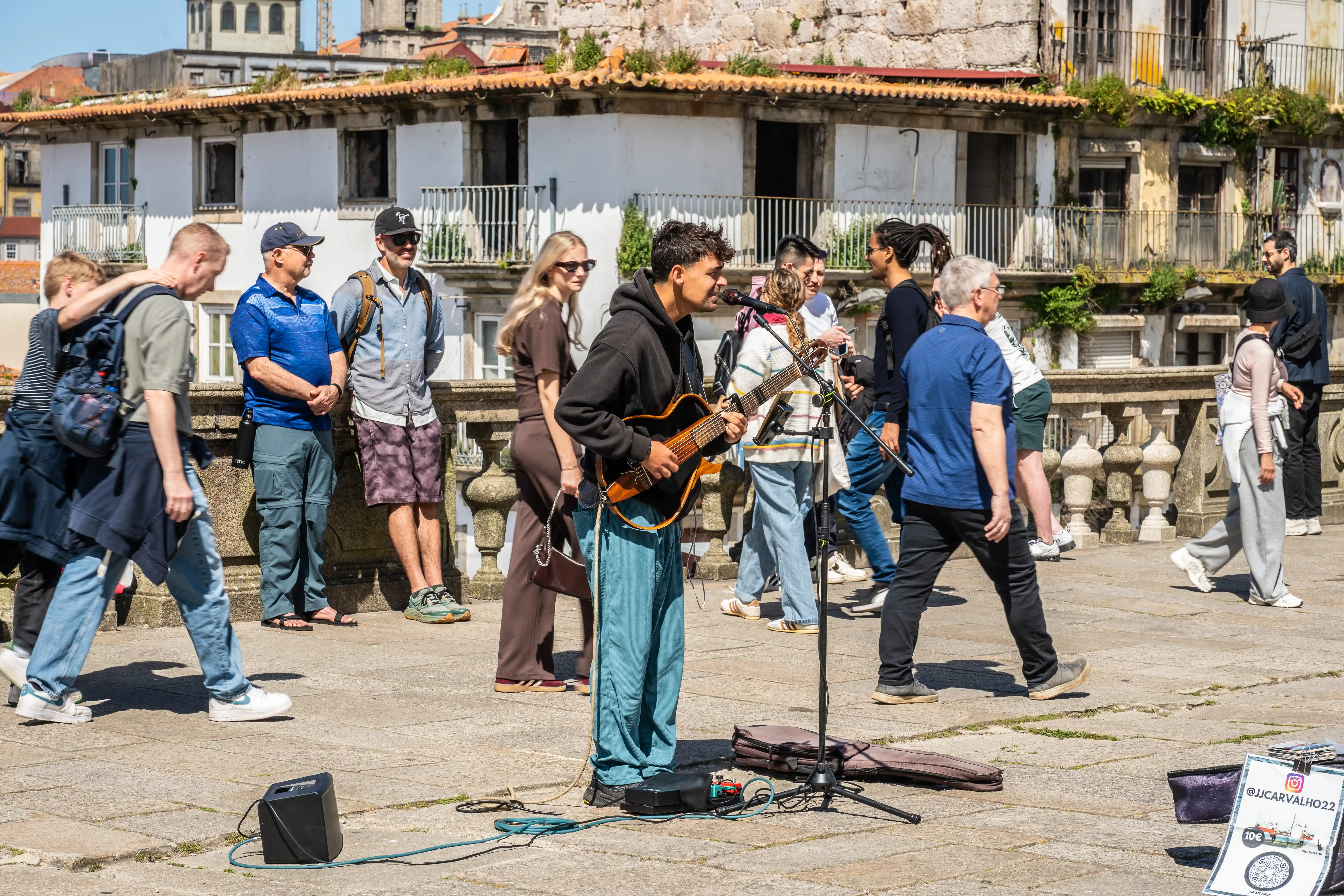  A street musician plays a guitar and sings into a microphone on a sunny day in a plaza, with a crowd of tourists watching.