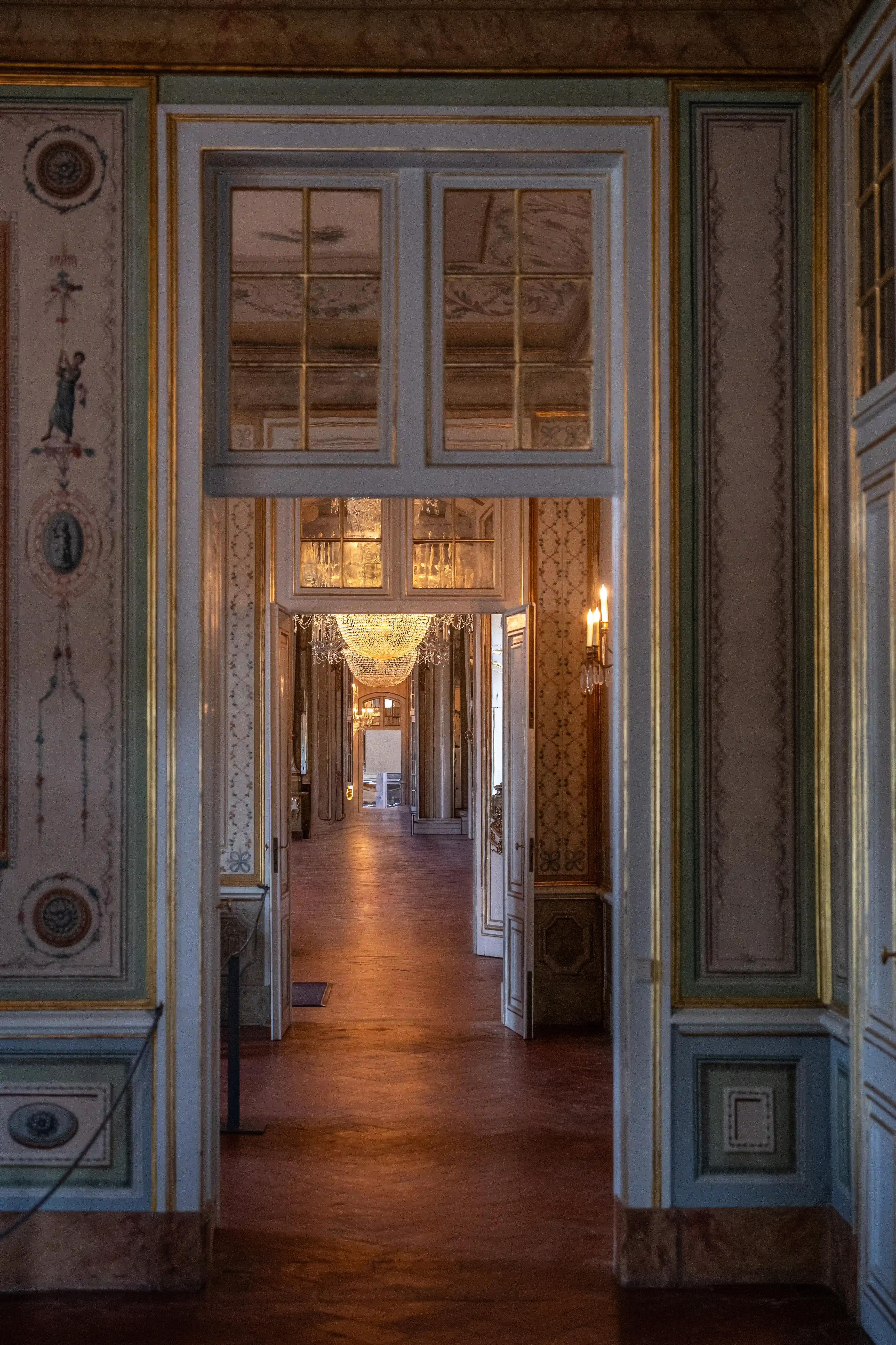 A long, opulent palace corridor with polished wooden floors. Several people are visible walking down the corridor. The walls are adorned with patterned wallpaper, decorative mouldings, and sconces. Large crystal chandeliers hang from the ceiling, and mirrored panels reflect the ornate decor.