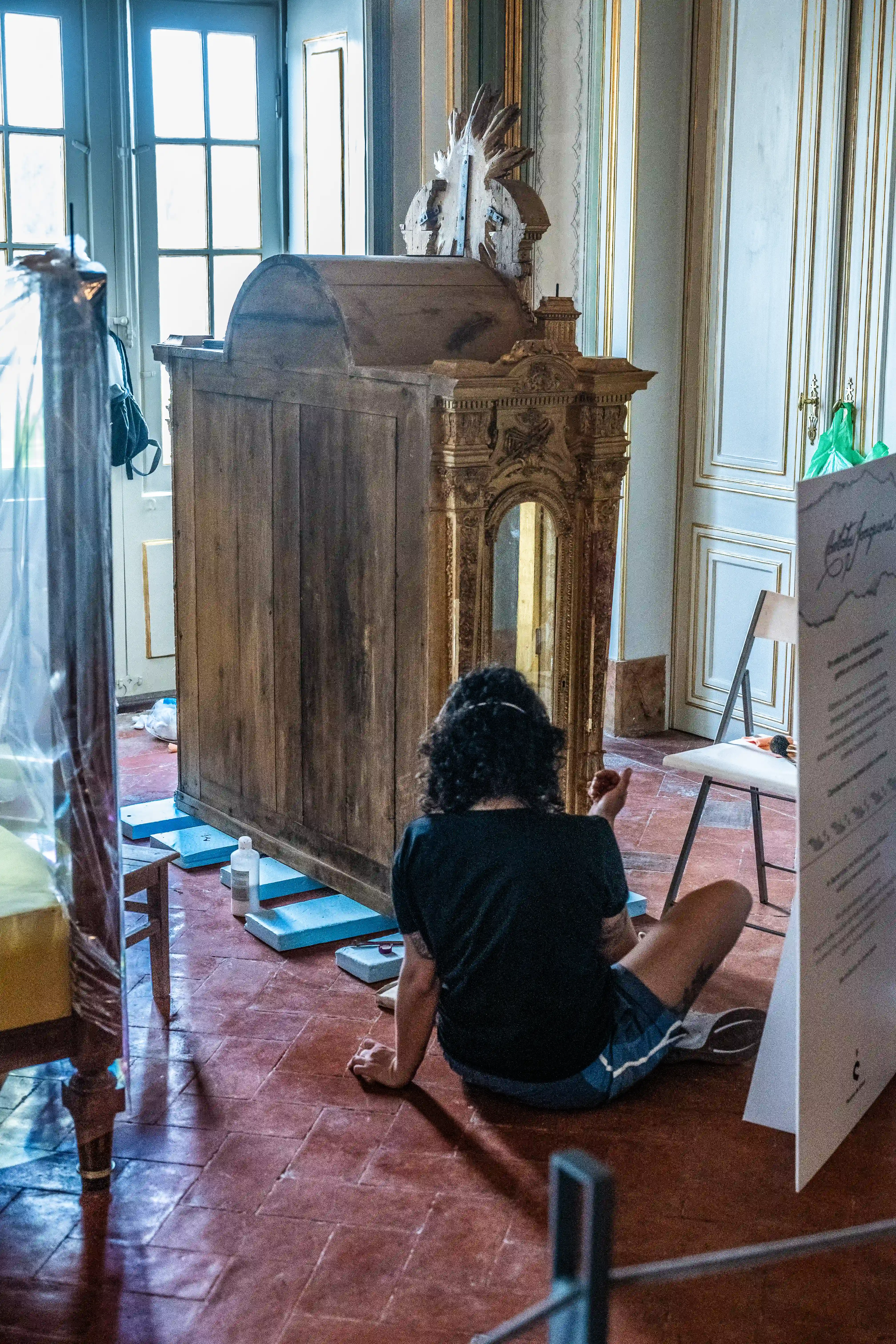  A person with curly dark hair sits on a terracotta tiled floor, facing away from the camera, working on a large, dark wood antique cabinet. The cabinet features ornate gilded embellishments and a mirrored opening. The room has elegant decorative wall panelling, large windows, and other furniture being protected.