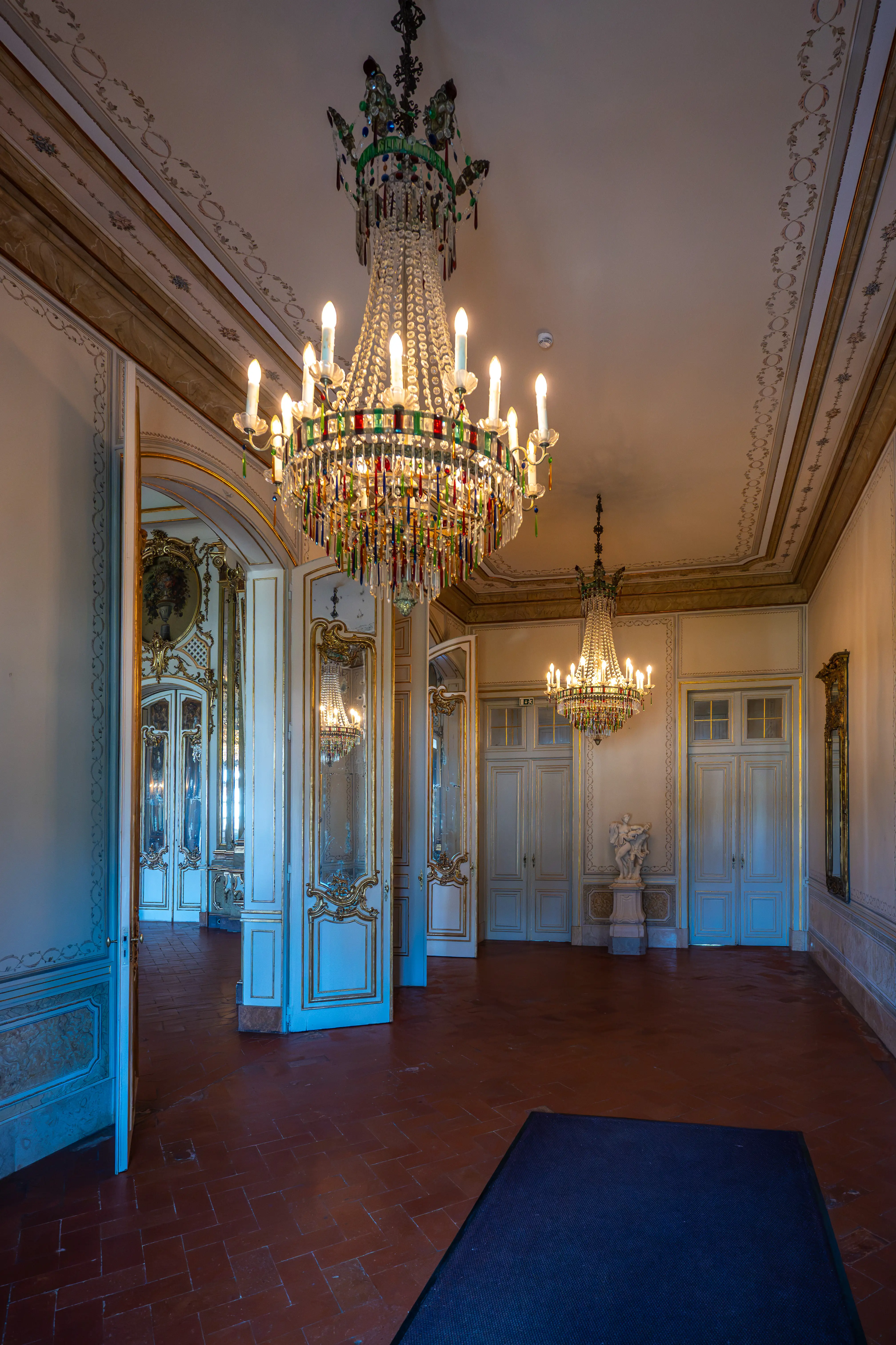 A dimly lit grand hall featuring two large, ornate crystal chandeliers. White doors with decorative gold trim and mirrored panels line the walls, reflecting the light from the chandeliers and the windows beyond.