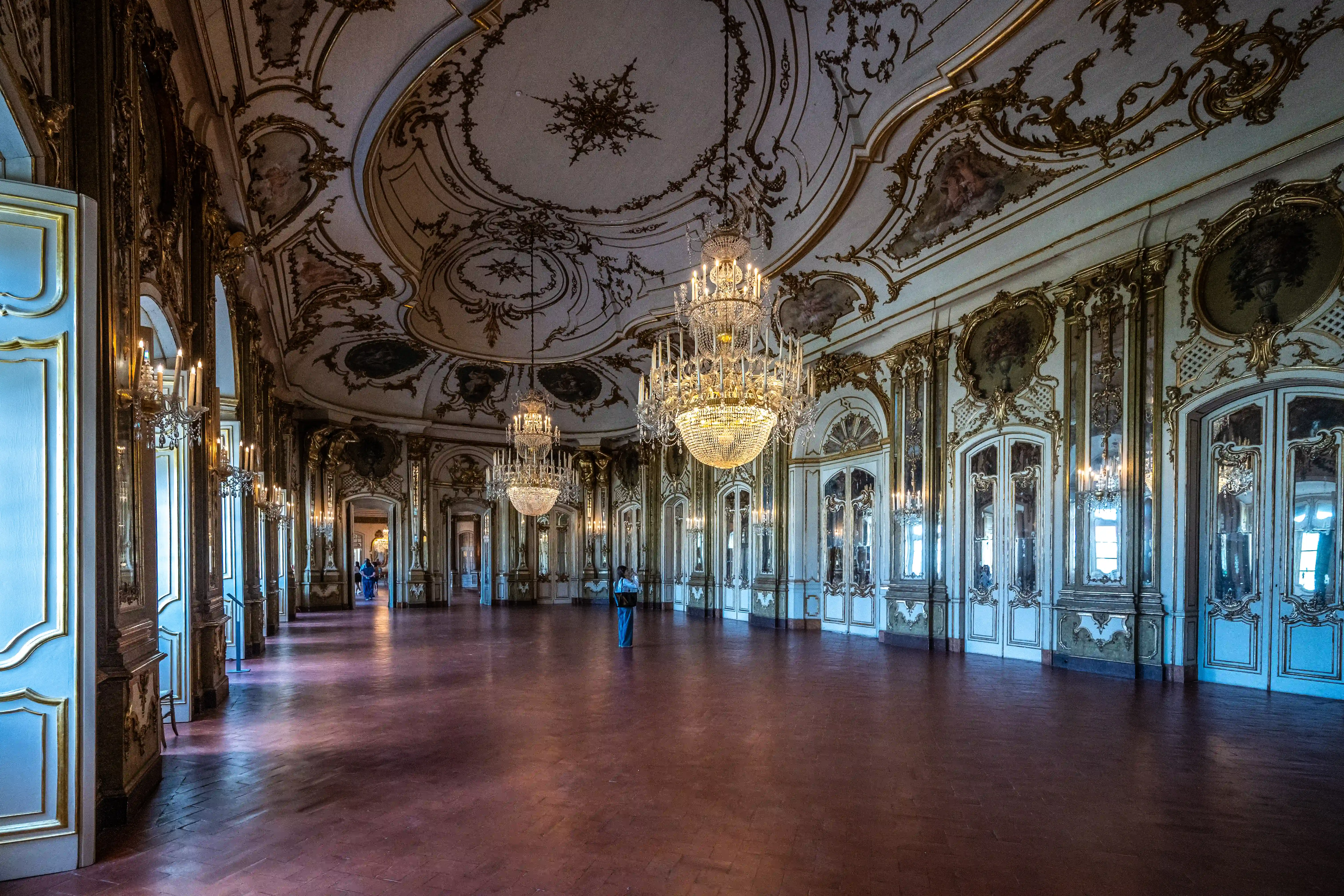 Interior view of a large, luxurious ballroom with a highly decorated ceiling, ornate gold trim on the walls, and large crystal chandeliers.