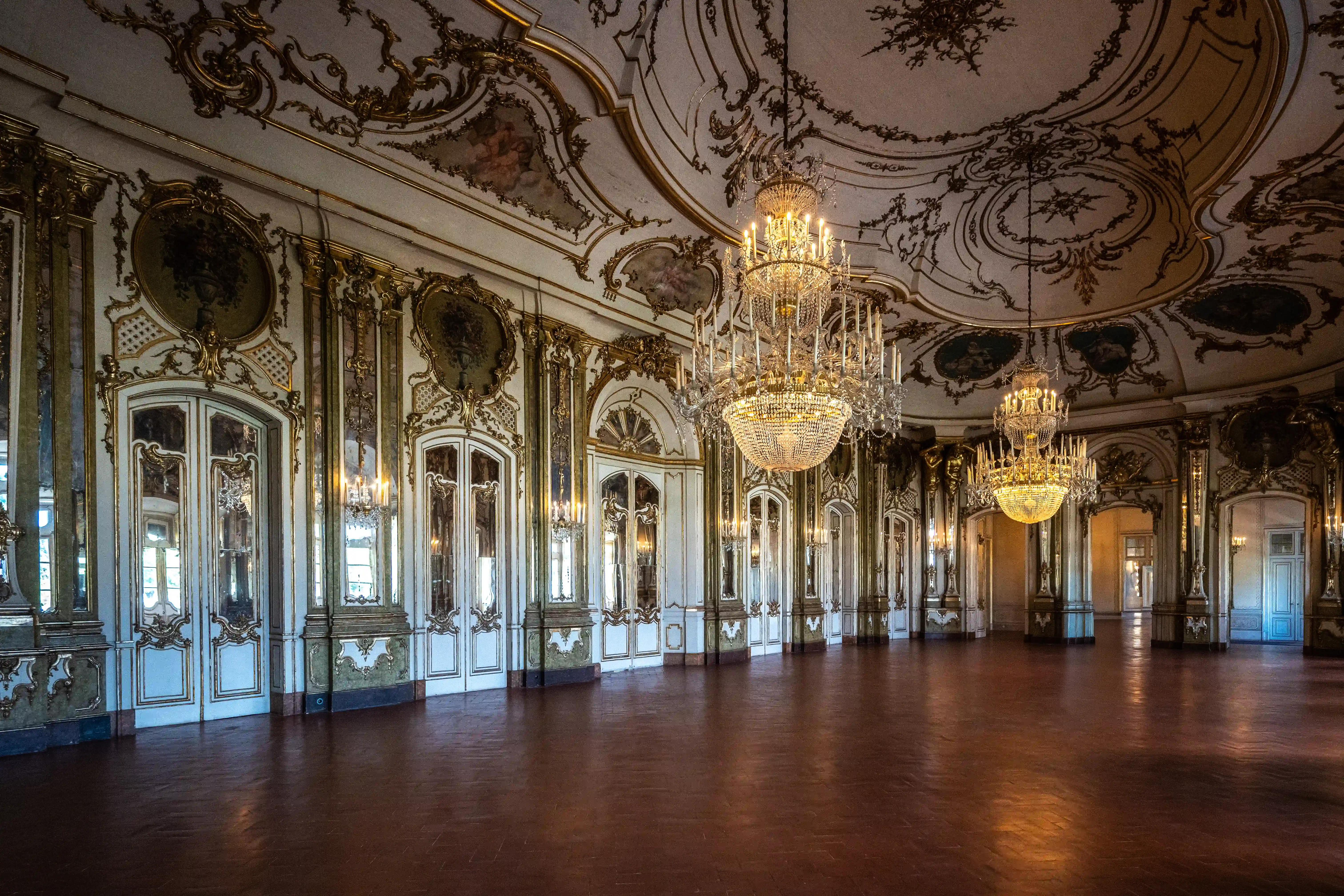 Wide shot of an empty, luxurious ballroom interior. The walls are decorated with ornate gold carvings and mirrored panels, featuring tall, white doors with decorative gold trim. Three large, multi-tiered crystal chandeliers hang from the elaborately painted ceiling. The floor is polished dark wood. A few figures can be seen in the distance, adding a sense of scale.