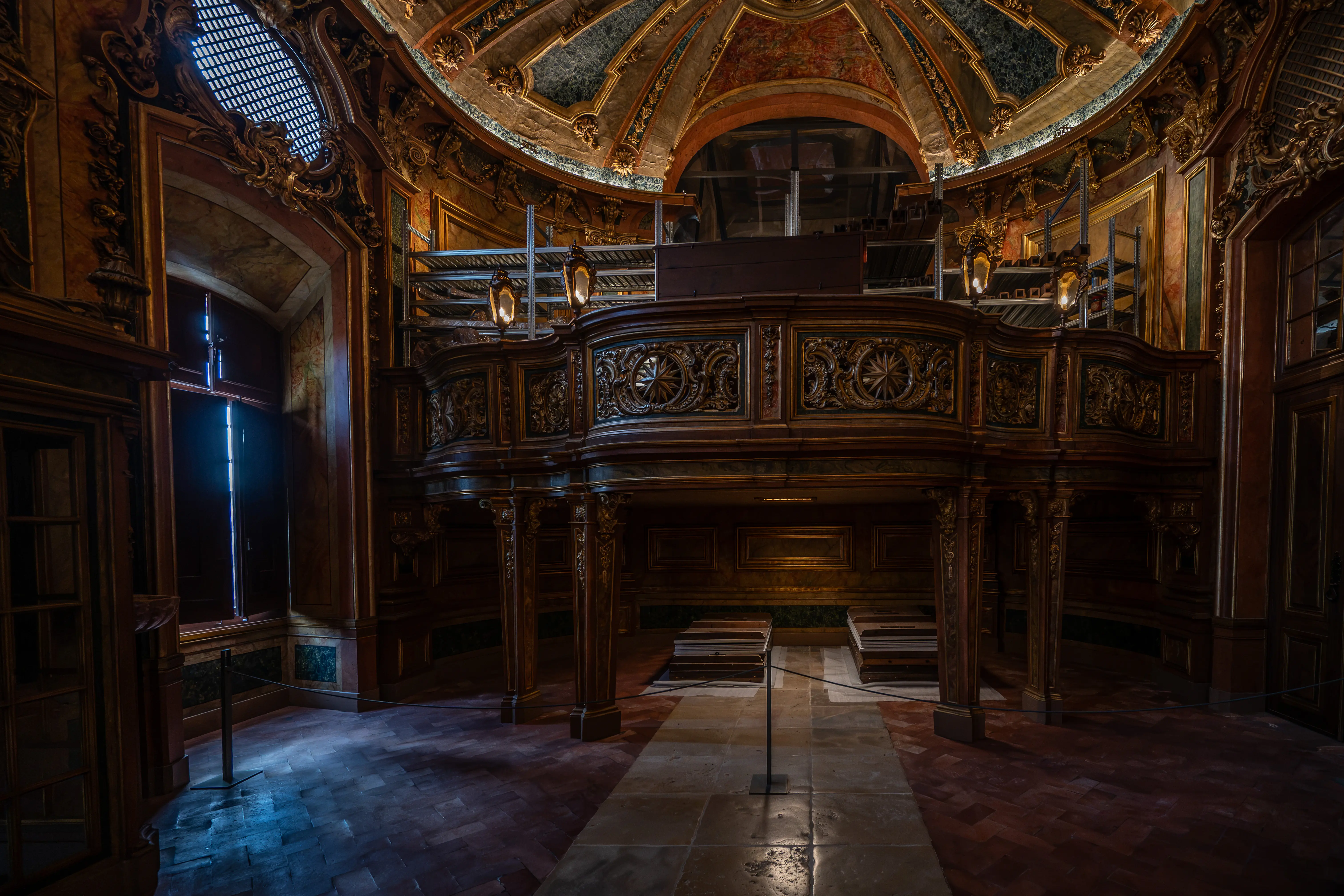 Interior view of a dark, ornately carved wooden organ loft in a Baroque building. Bright light from a high window illuminates the floor