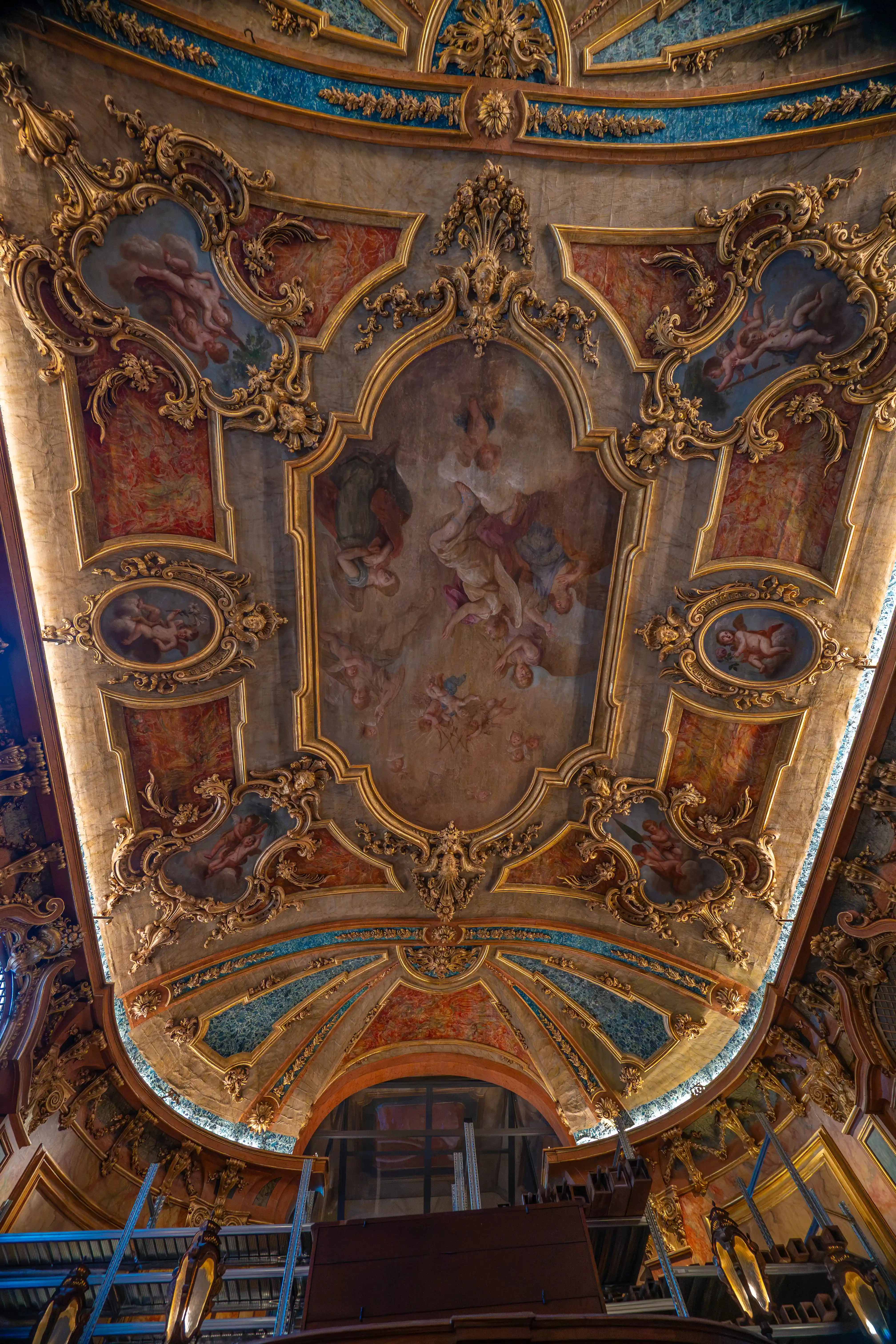 A low-angle shot of an elaborately decorated ceiling in a Baroque style, with gilded mouldings surrounding painted panels showing figures in the sky.