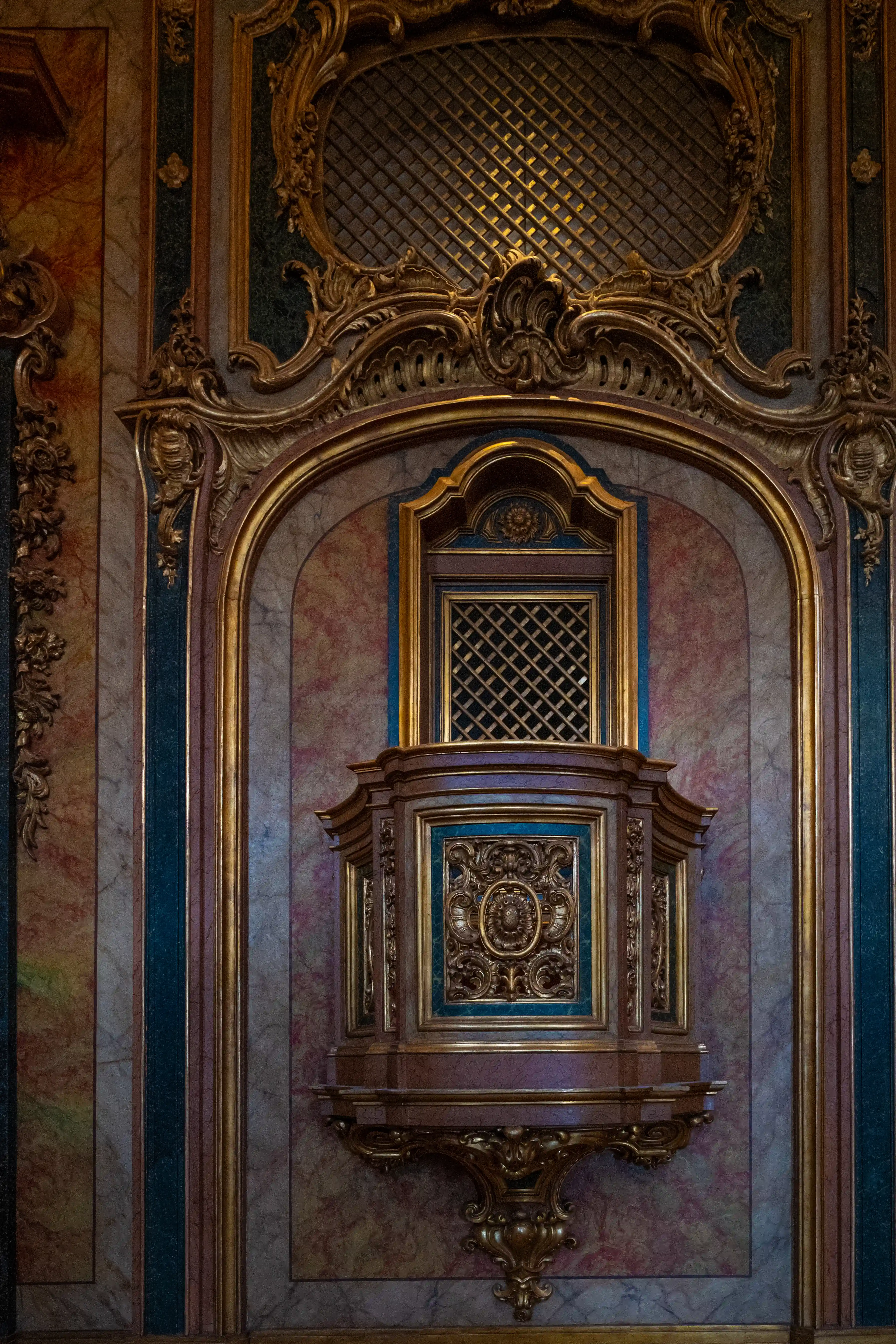 A close-up view of a dark, ornate pulpit with gilded carvings and a patterned screen, set against a marble-patterned wall with decorative gold and dark green trim.