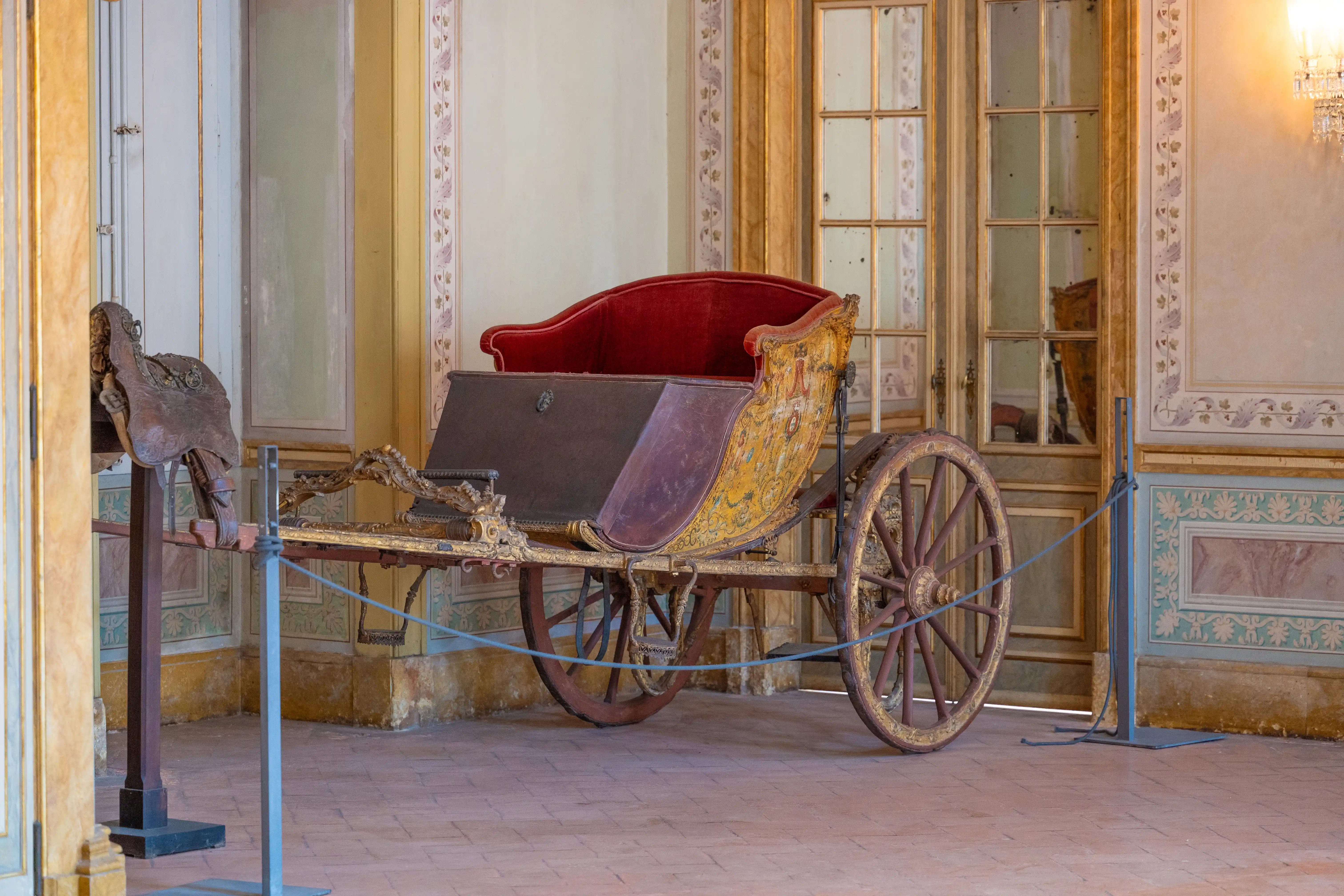 A vintage carriage with red upholstery and intricate gold decoration, resting on wooden wheels, is showcased indoors.