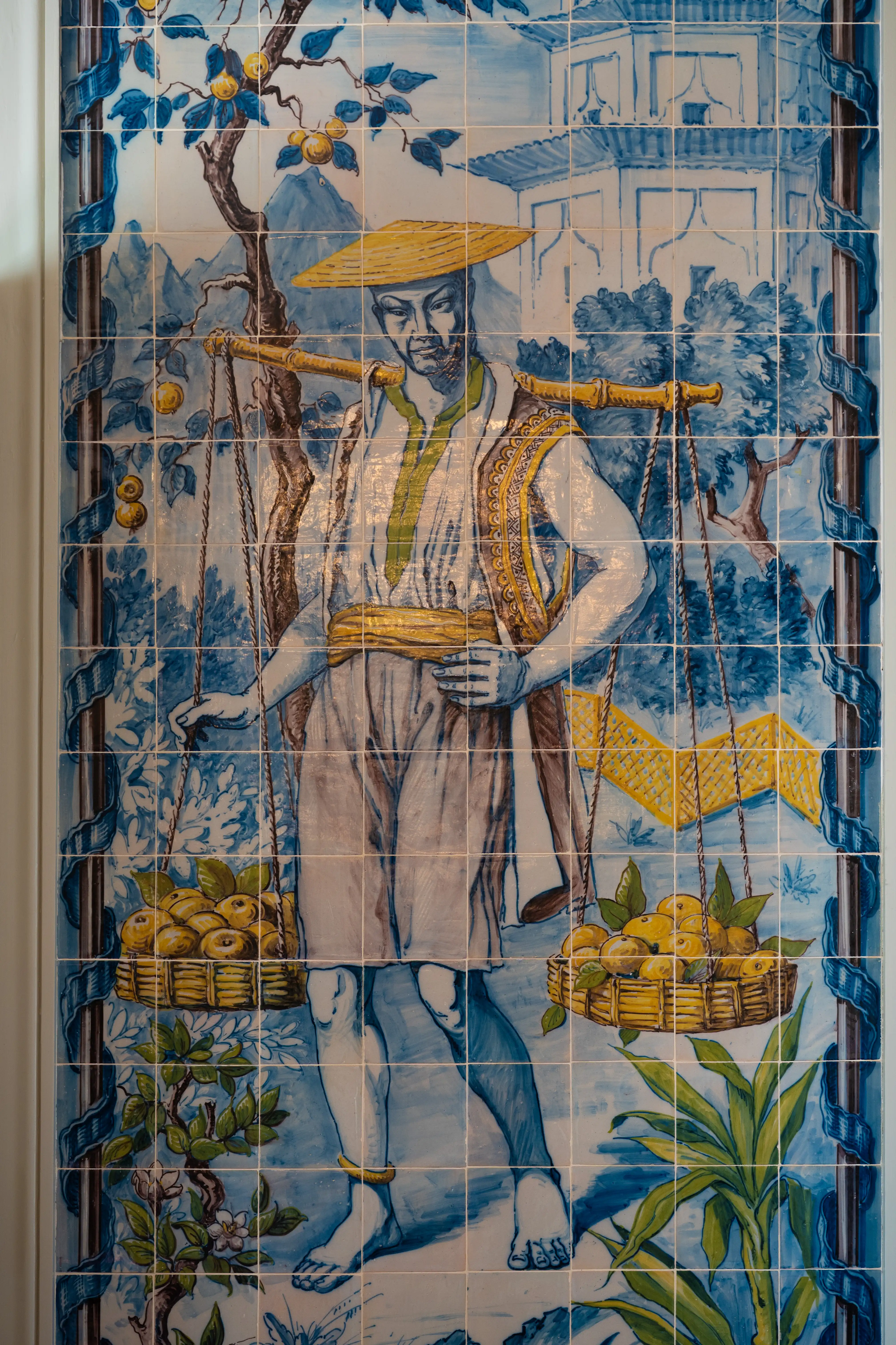 Ceramic tile art of a person carrying baskets of fruit on a yoke. The person wears a straw hat and traditional clothing, set against a blue and white landscape with trees and buildings.