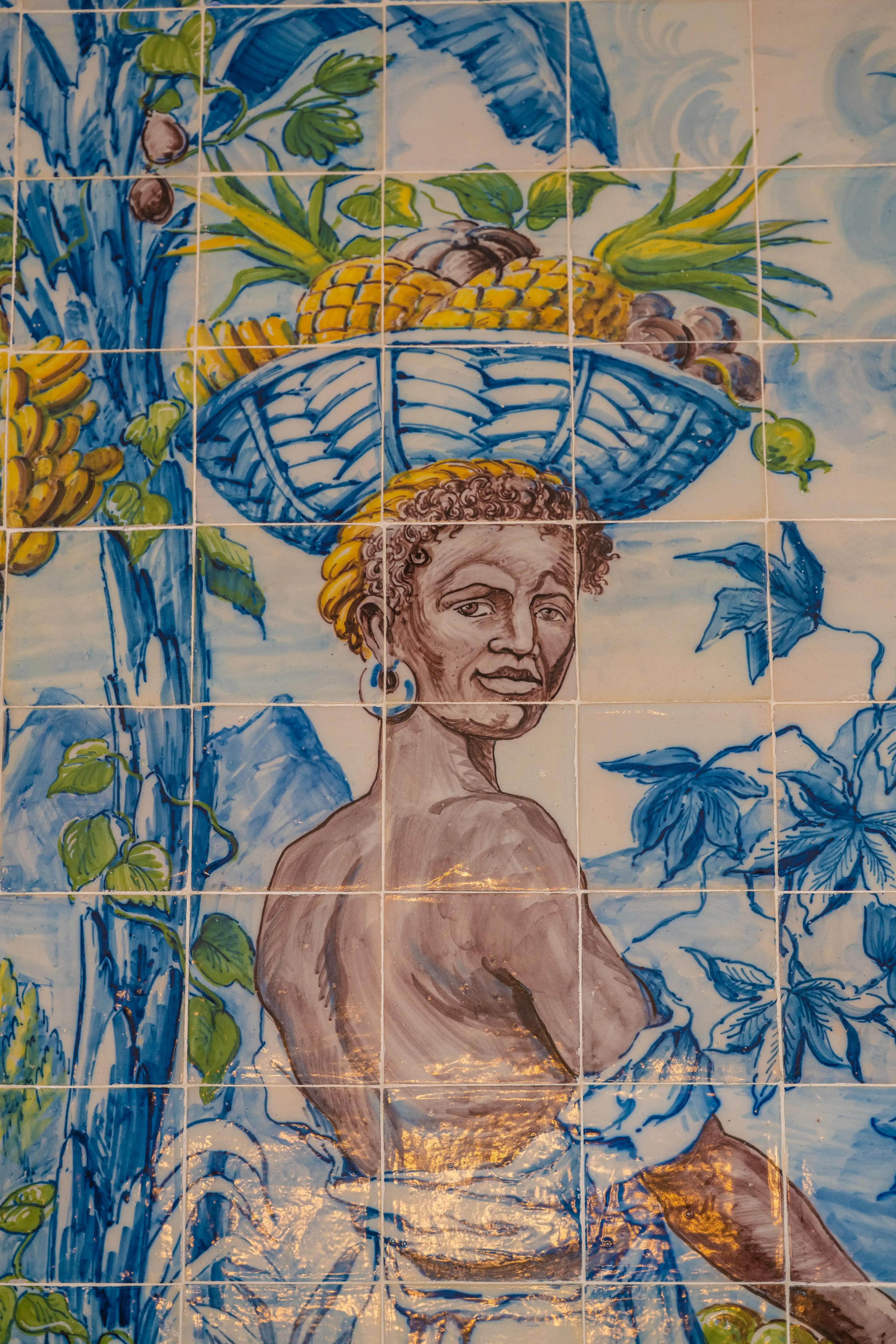 A close-up view of a large, rectangular azulejo tile panel featuring an allegorical scene in blue and white. A central female figure, wearing a hat and ornate clothing, stands beneath a tree. A cherubic figure is seated at her feet near a golden object. The panel is bordered by decorative elements and is next to a white wall with gilded trim and a window reflection.