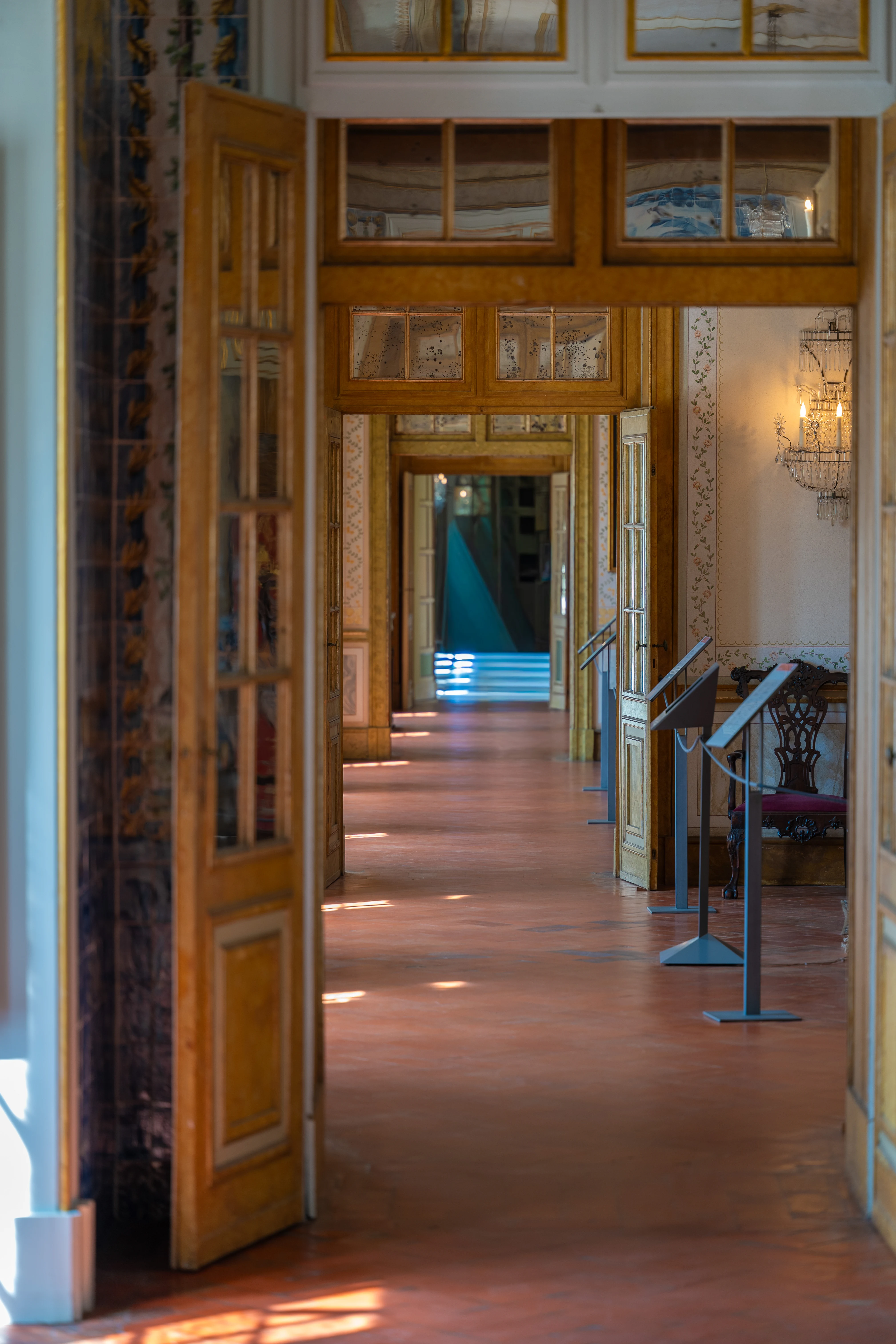 Interior view of a long, sunlit hallway with multiple open doorways. The floor is terracotta and reflects geometric patterns of light and shadow. Ornate wooden door frames, decorative wallpaper, and a chandelier are visible. Two people are seen walking in the distance