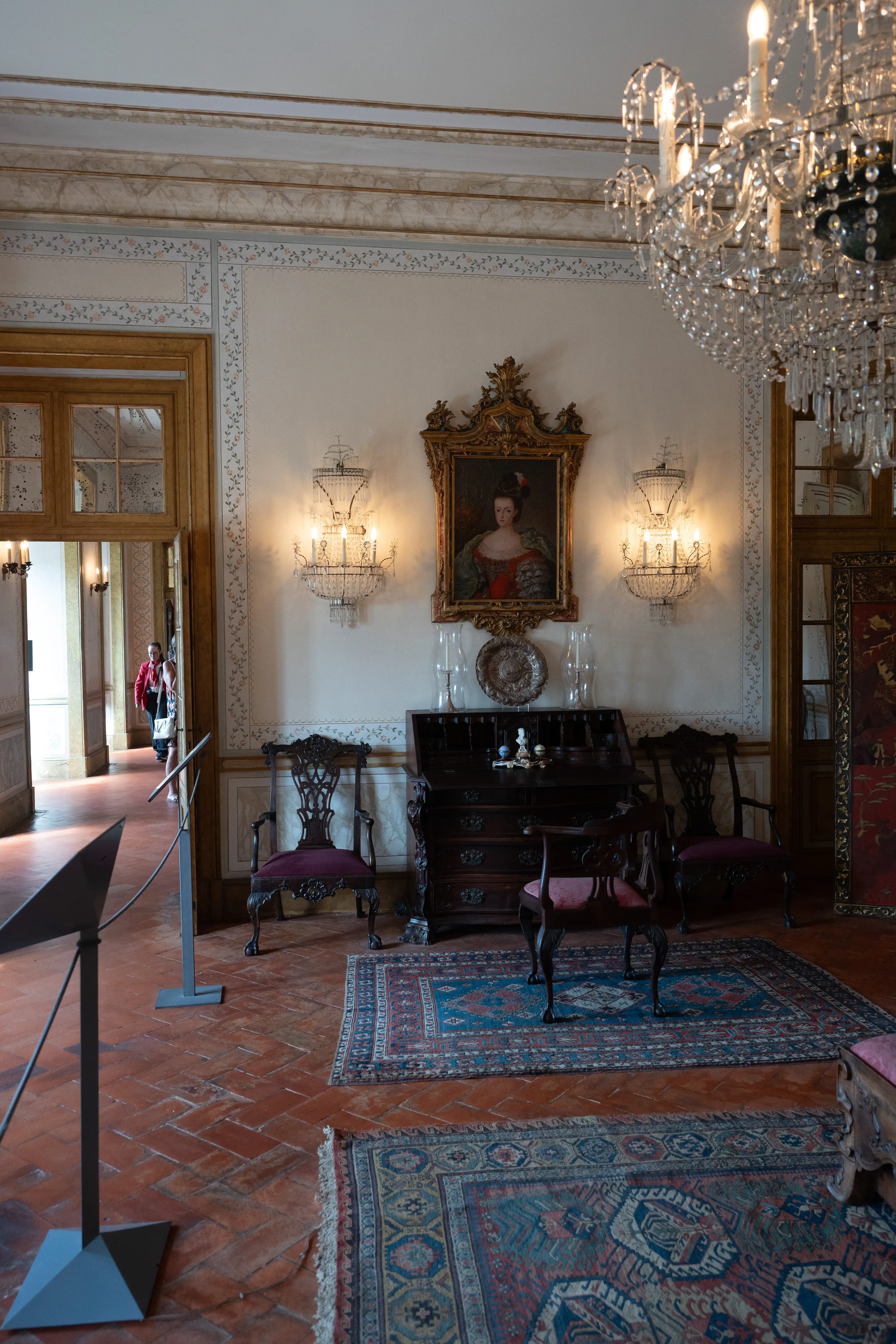  Interior view of a long, sunlit hallway with multiple open doorways. The floor is terracotta and reflects geometric patterns of light and shadow. Ornate wooden door frames, decorative wallpaper, and a chandelier are visible. Two people are seen walking in the distance.