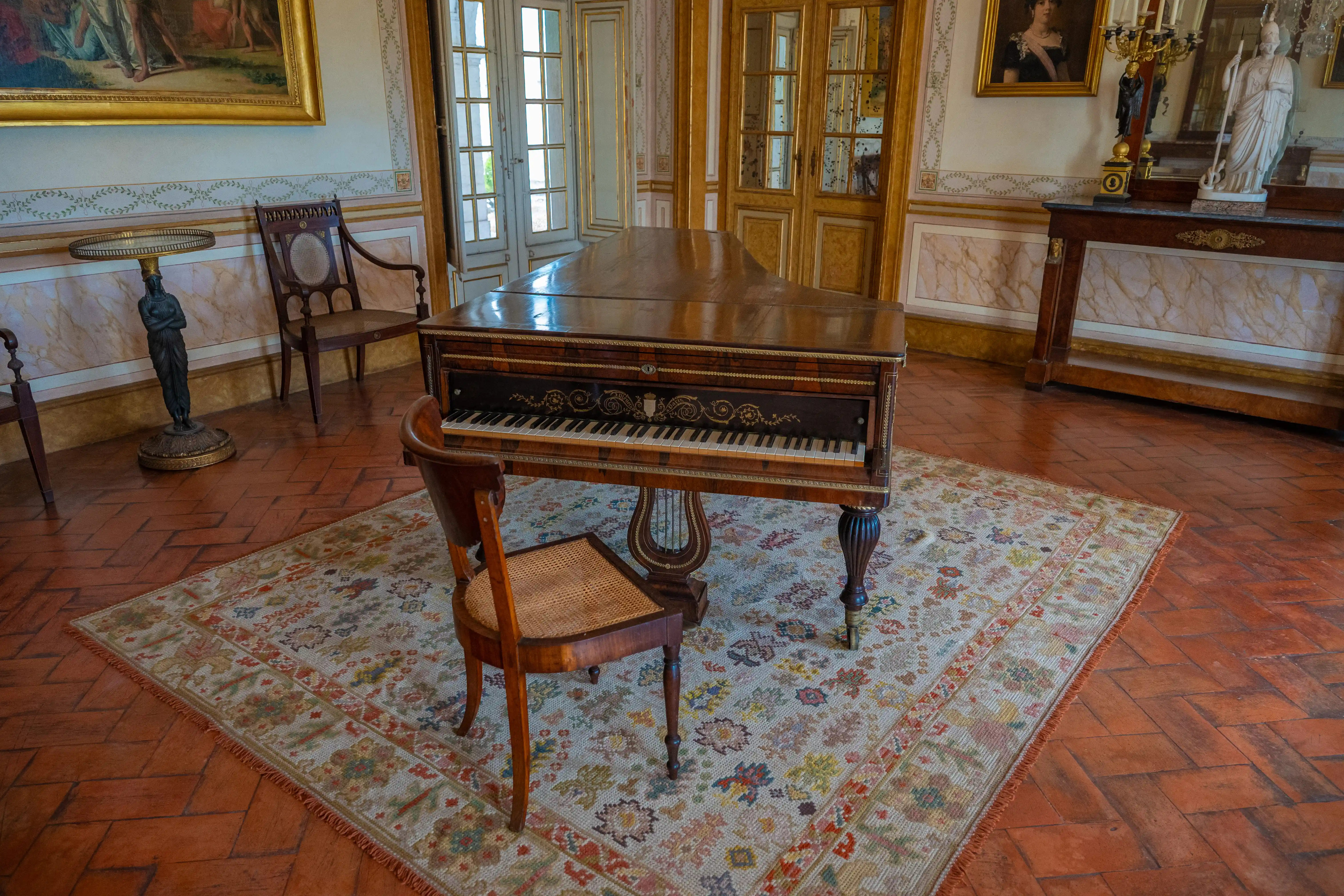 A polished wooden grand piano with ivory and ebony keys is positioned on a patterned rug in a stately room. A wooden chair with a woven seat is placed in front of the piano.