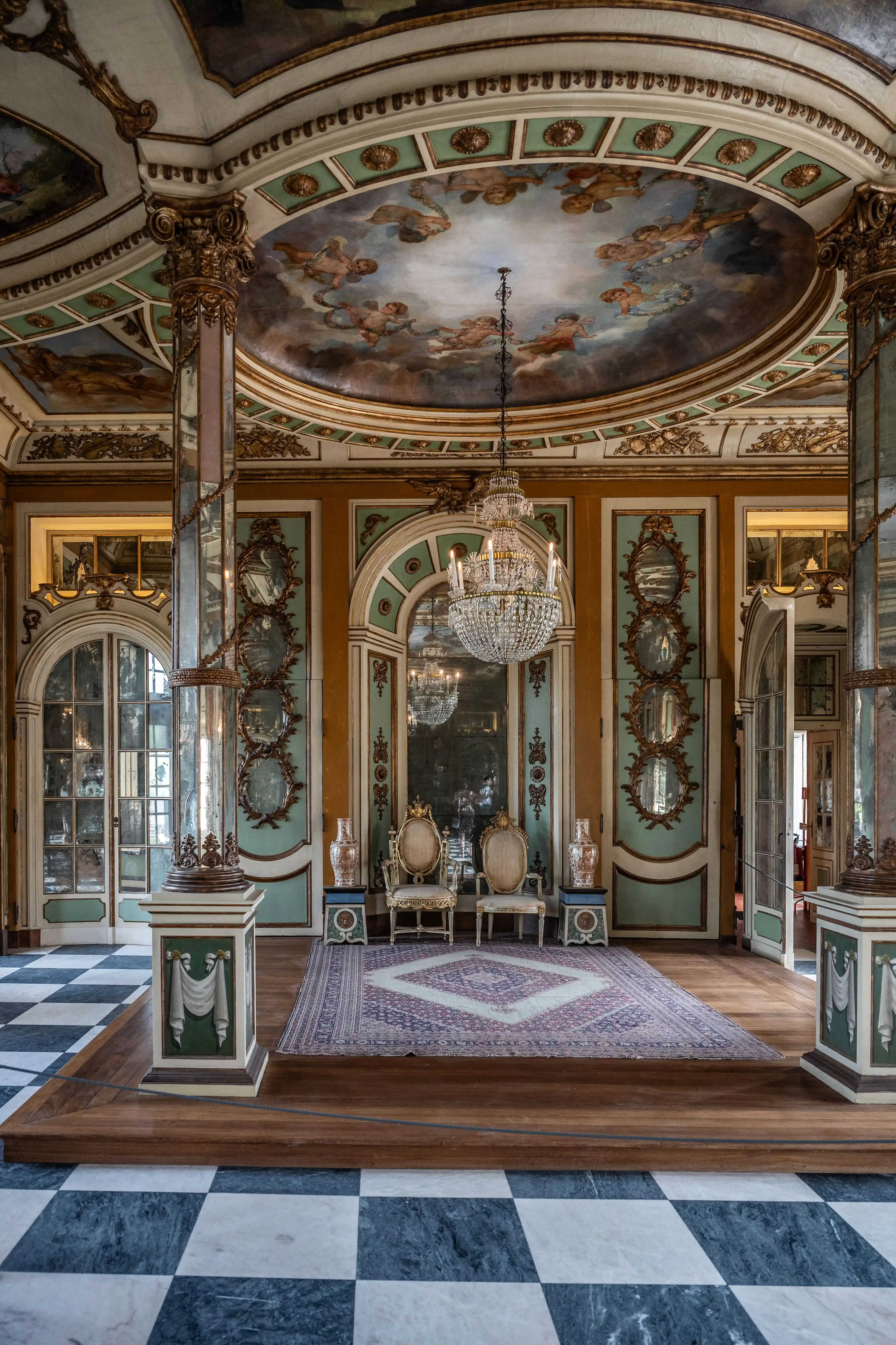 Interior view of a lavish Rococo salon. A large crystal chandelier hangs from a circular ceiling painted with cherubs. Two ornate gilded chairs sit on a raised wooden platform with a patterned rug. The walls are decorated with mint green panels, gold mouldings, and mirrored oval frames. The floor is a black and white checkerboard pattern.