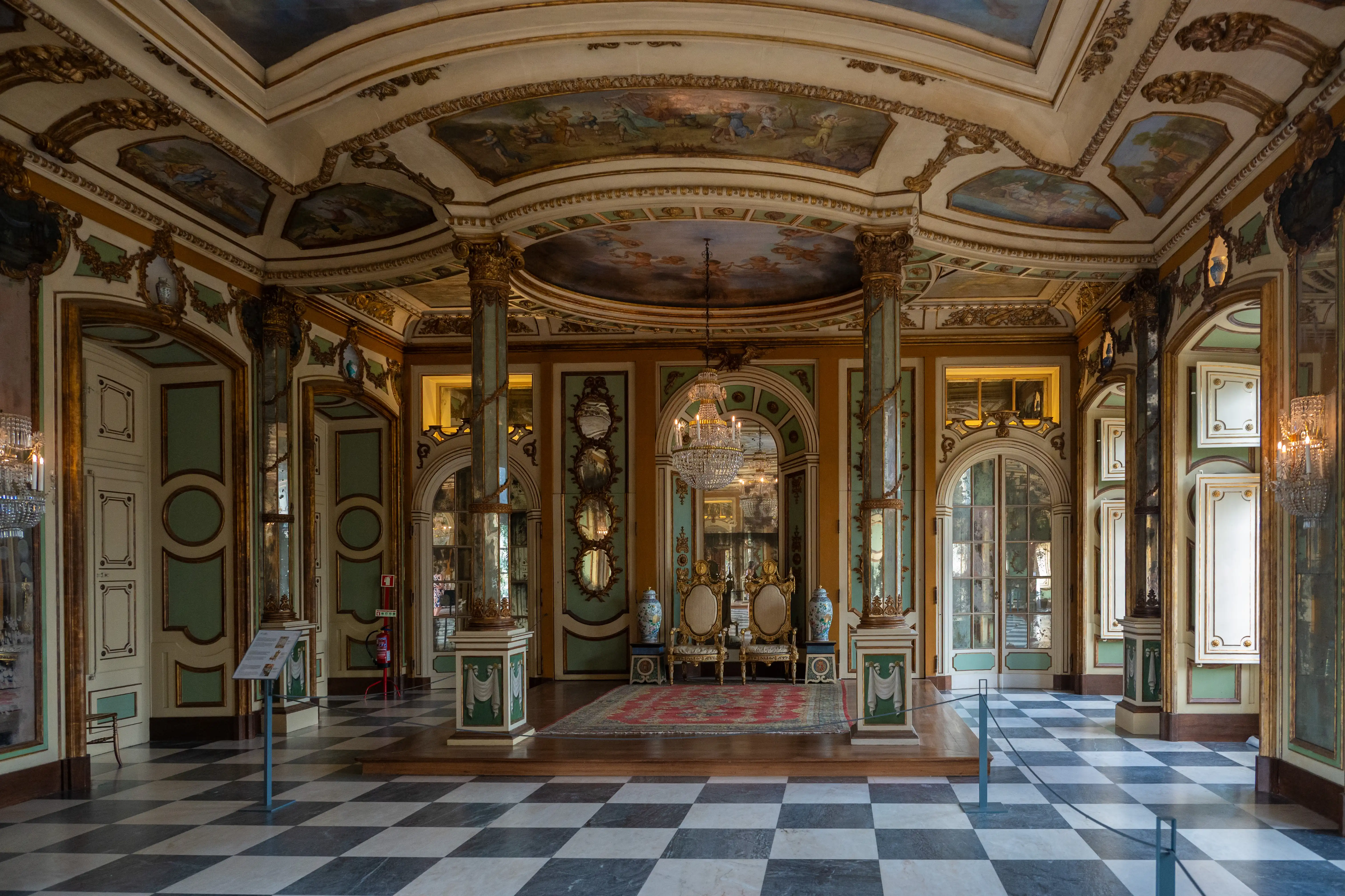 Interior view of a richly decorated ballroom featuring a checkerboard floor, gilded architectural details, painted ceiling frescoes, mirrored walls, and a central crystal chandelier.