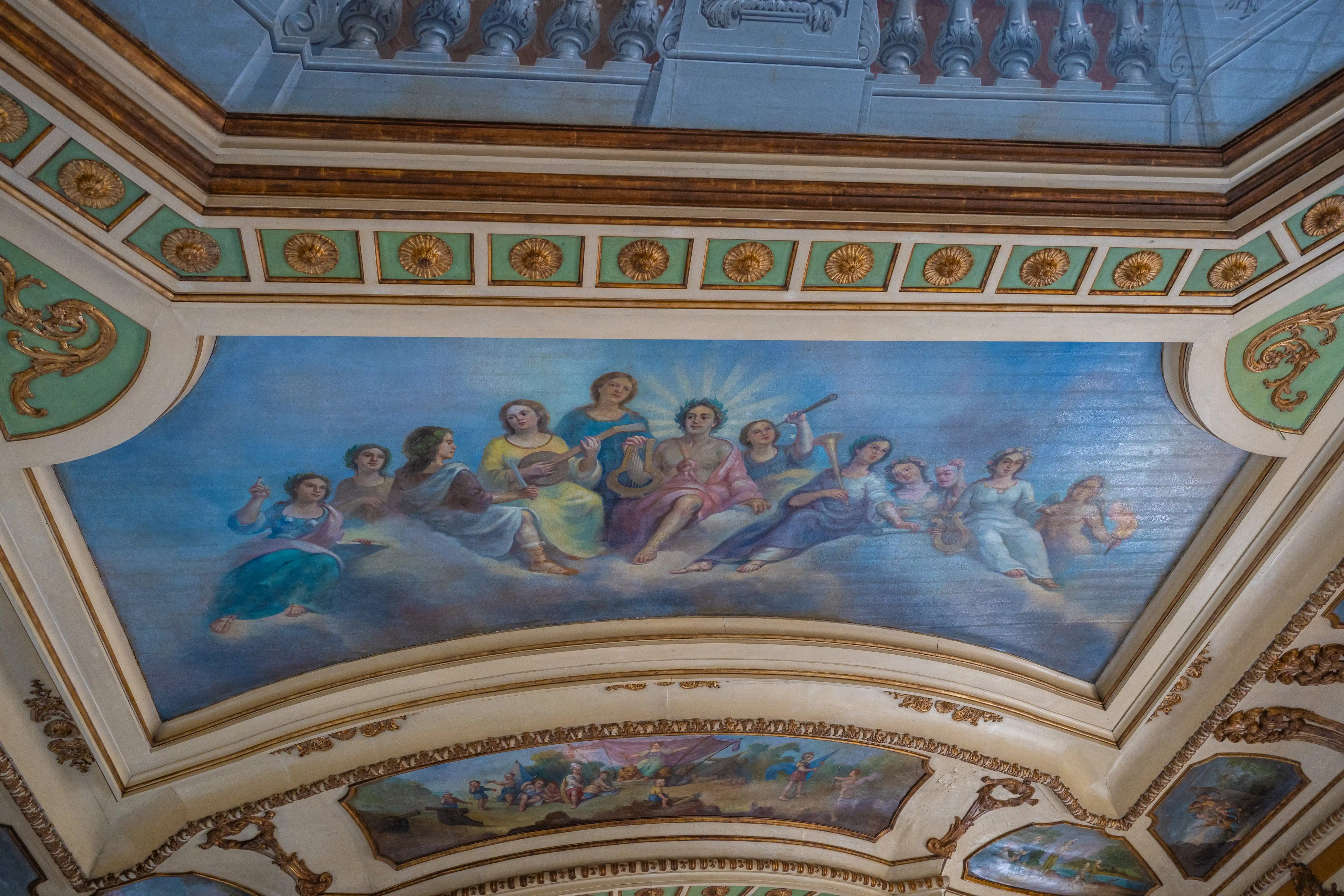 A ceiling fresco shows a group of figures, some playing instruments like lutes and horns, floating on clouds under a light blue sky. Decorative mouldings surround the fresco.