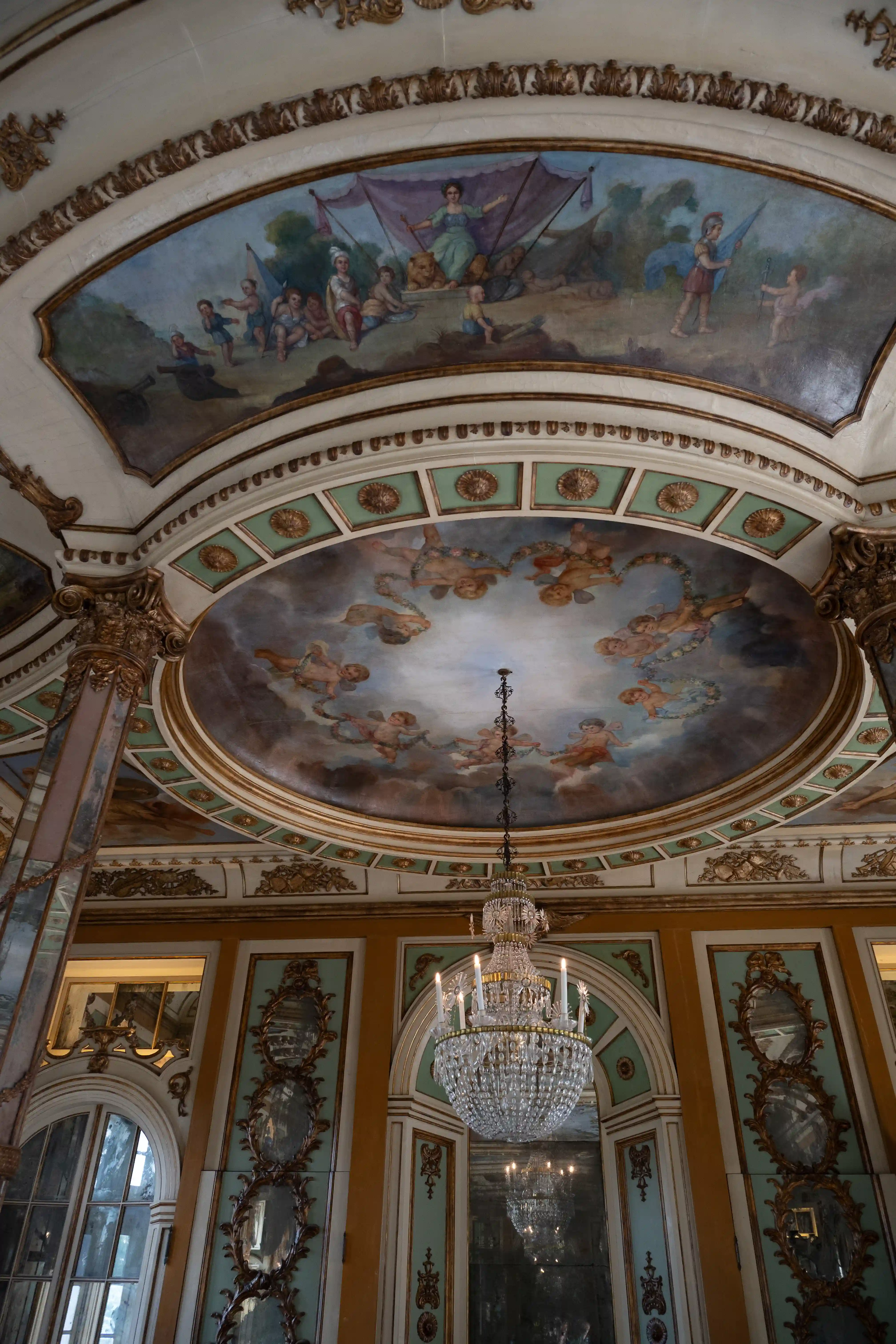 A low-angle view looking up at a highly decorative ceiling with painted frescoes of cherubs and floral elements, surrounded by gilded architectural details and columns.