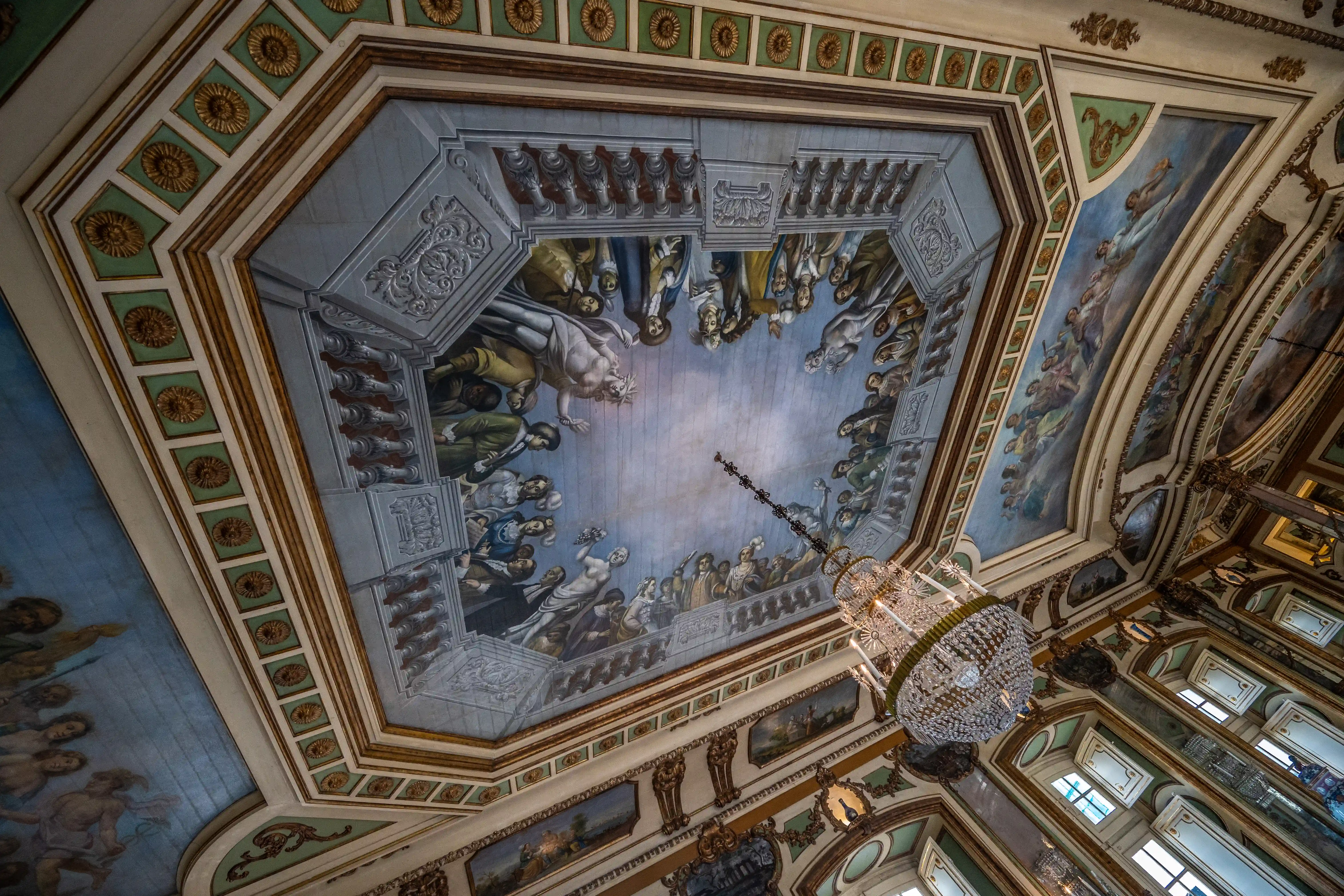 A low-angle view looking up at a highly decorative ceiling with painted frescoes of cherubs and floral elements, surrounded by gilded architectural details and columns.