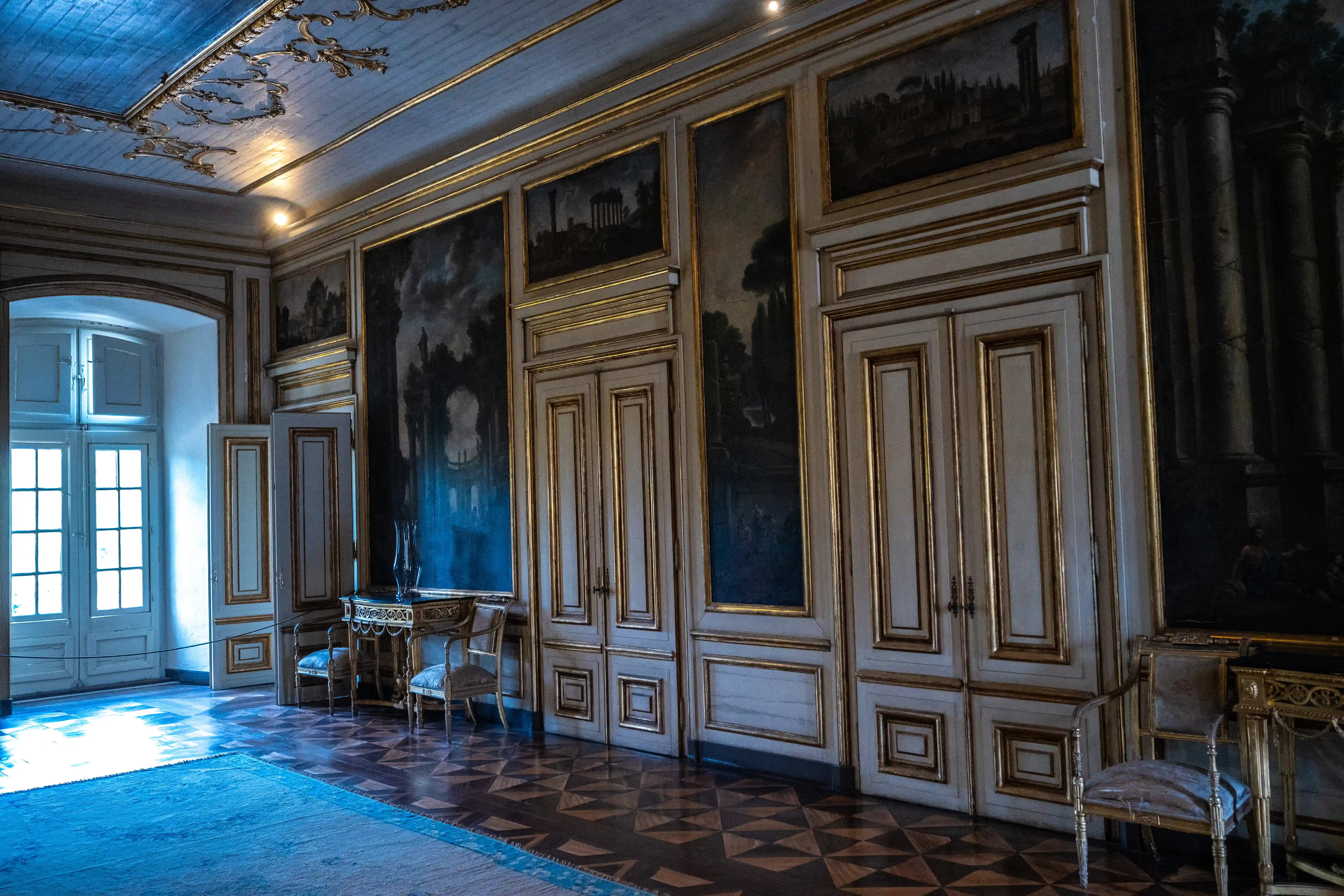  Interior view of a richly decorated salon with frescoed walls, gold mouldings, double doors, antique furniture, and a patterned wooden floor.
