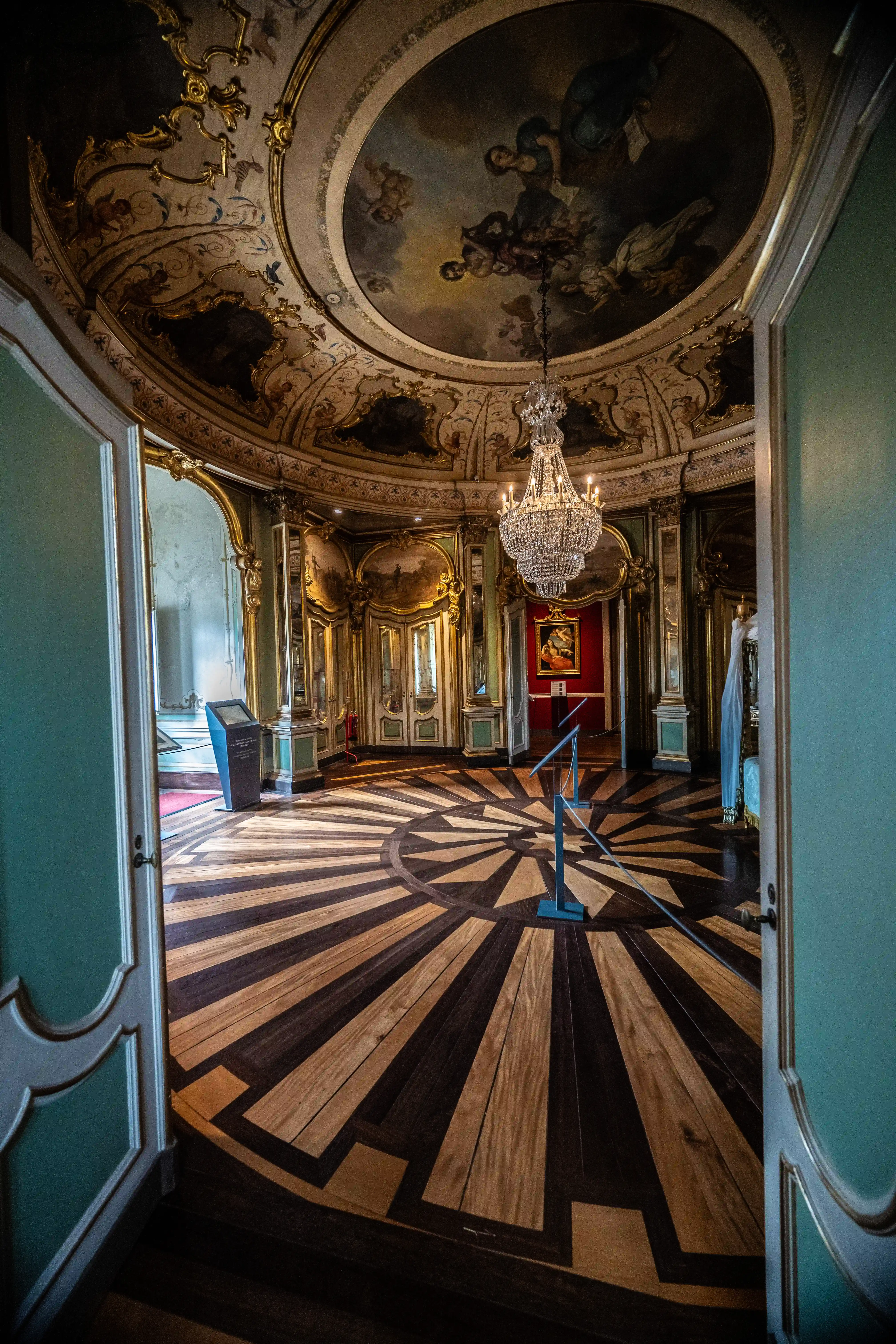 Interior view of a grand salon, showcasing a circular frescoed ceiling, intricate gold decorations, a large crystal chandelier, and a detailed sunburst wooden floor.