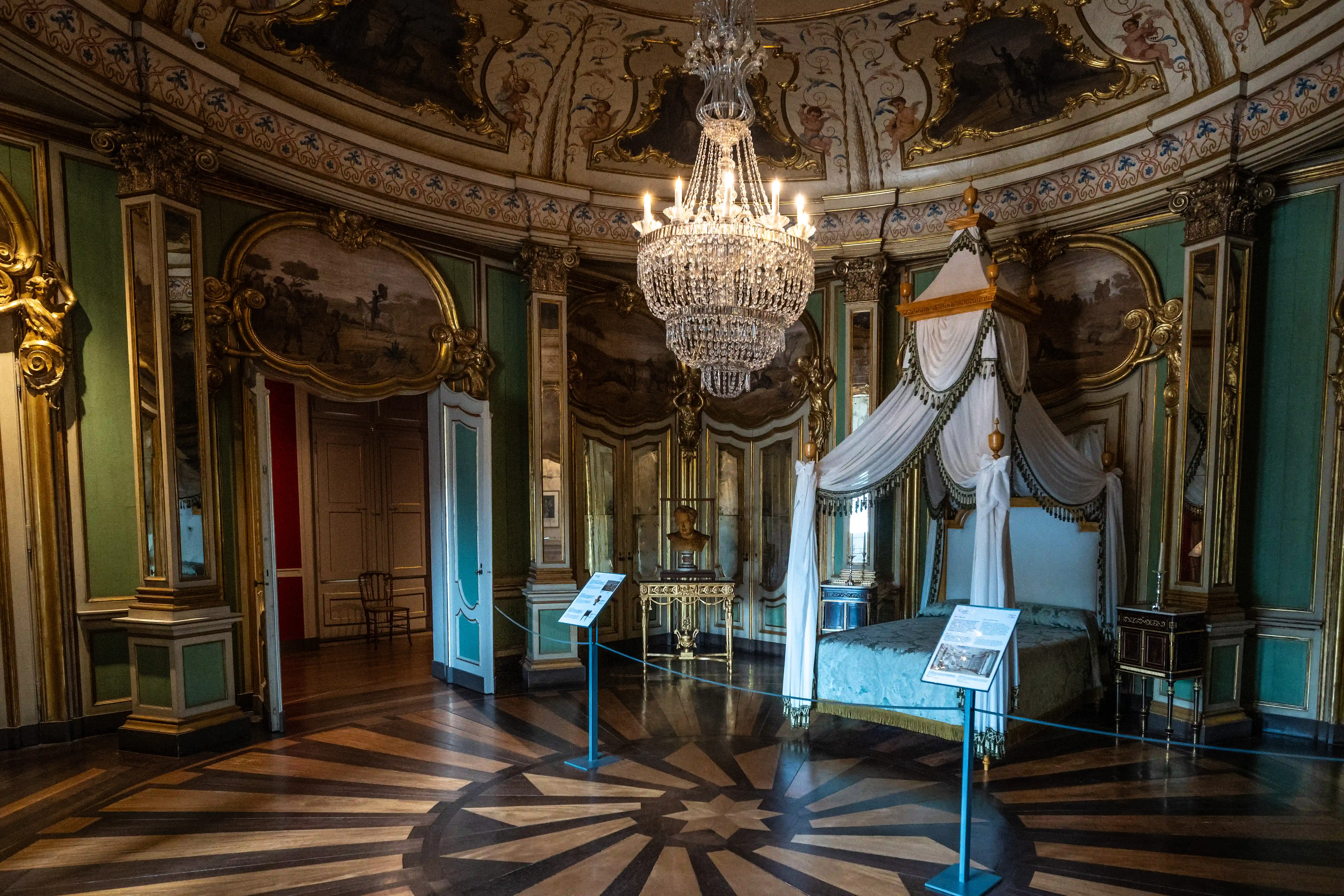 A wide shot of a richly decorated bedroom, showing a large canopy bed, a sparkling crystal chandelier, ornate wall panels, and a patterned wooden floor.