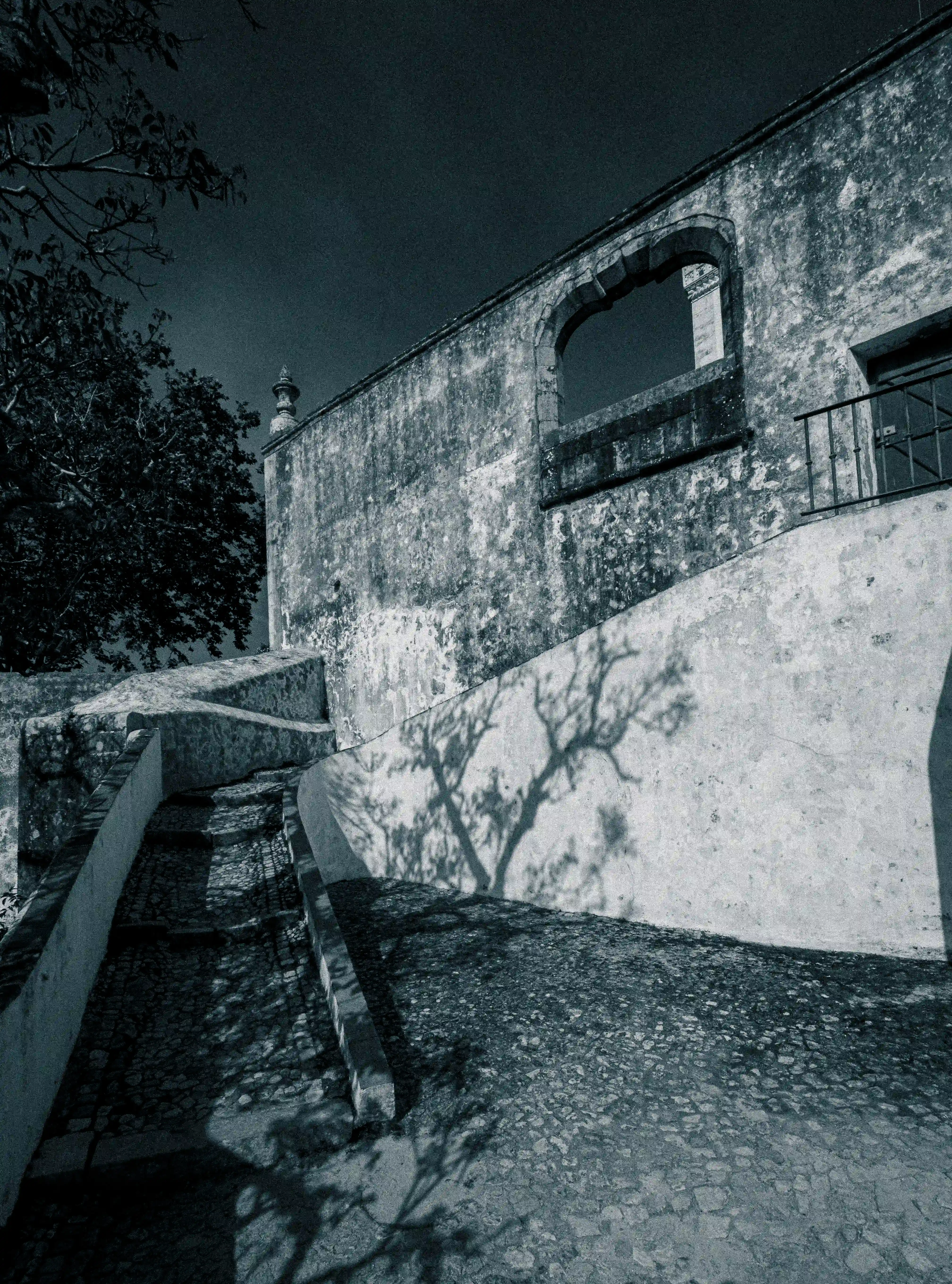Worn cobblestone steps with white and terracotta edging lead up a sloped path towards an old stone building. The building has a large, open archway and a wooden door with a balcony. Shadows of tree branches are cast across the white walls.