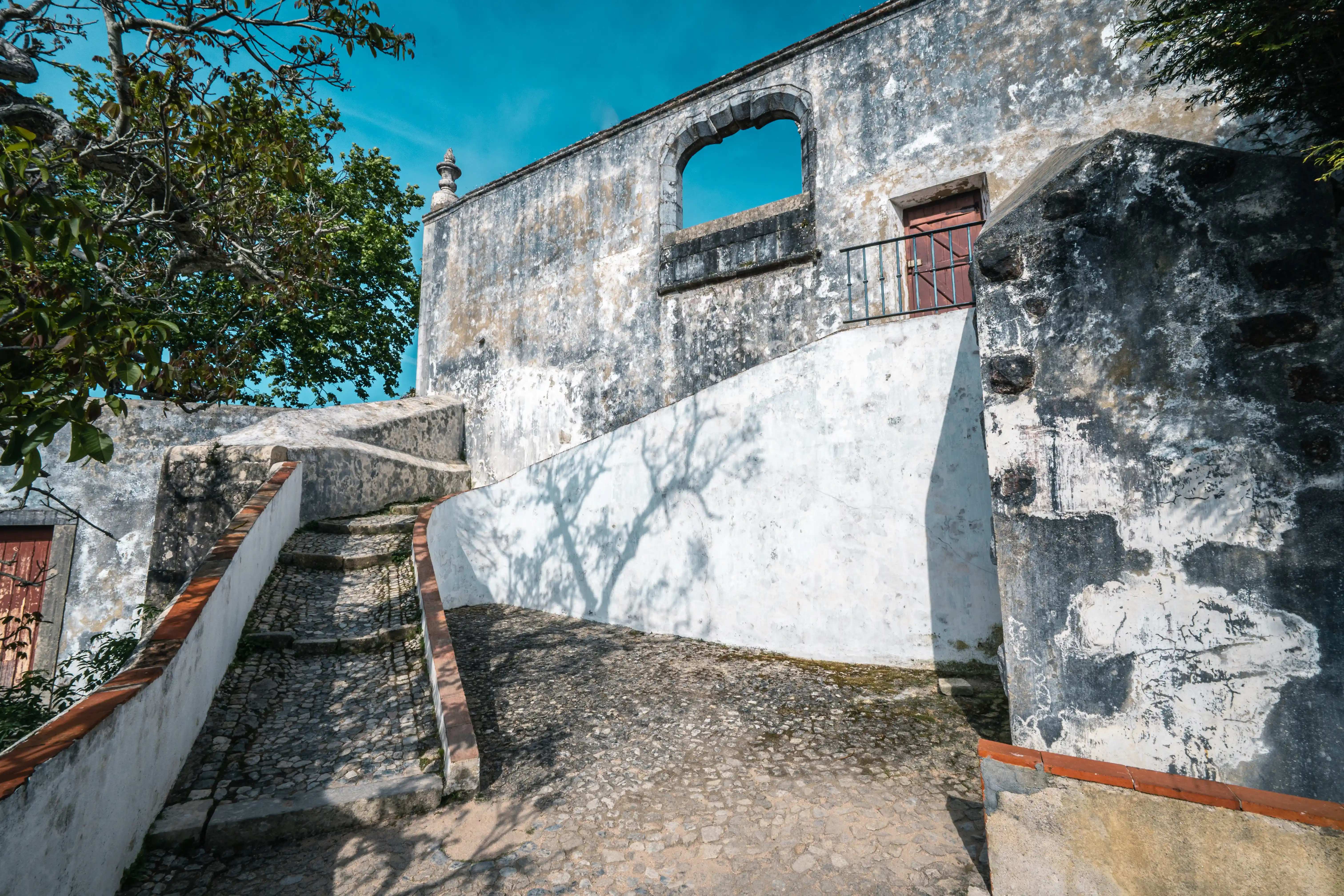 Worn cobblestone steps with white and terracotta edging lead up a sloped path towards an old stone building. The building has a large, open archway and a wooden door with a balcony. Shadows of tree branches are cast across the white walls.