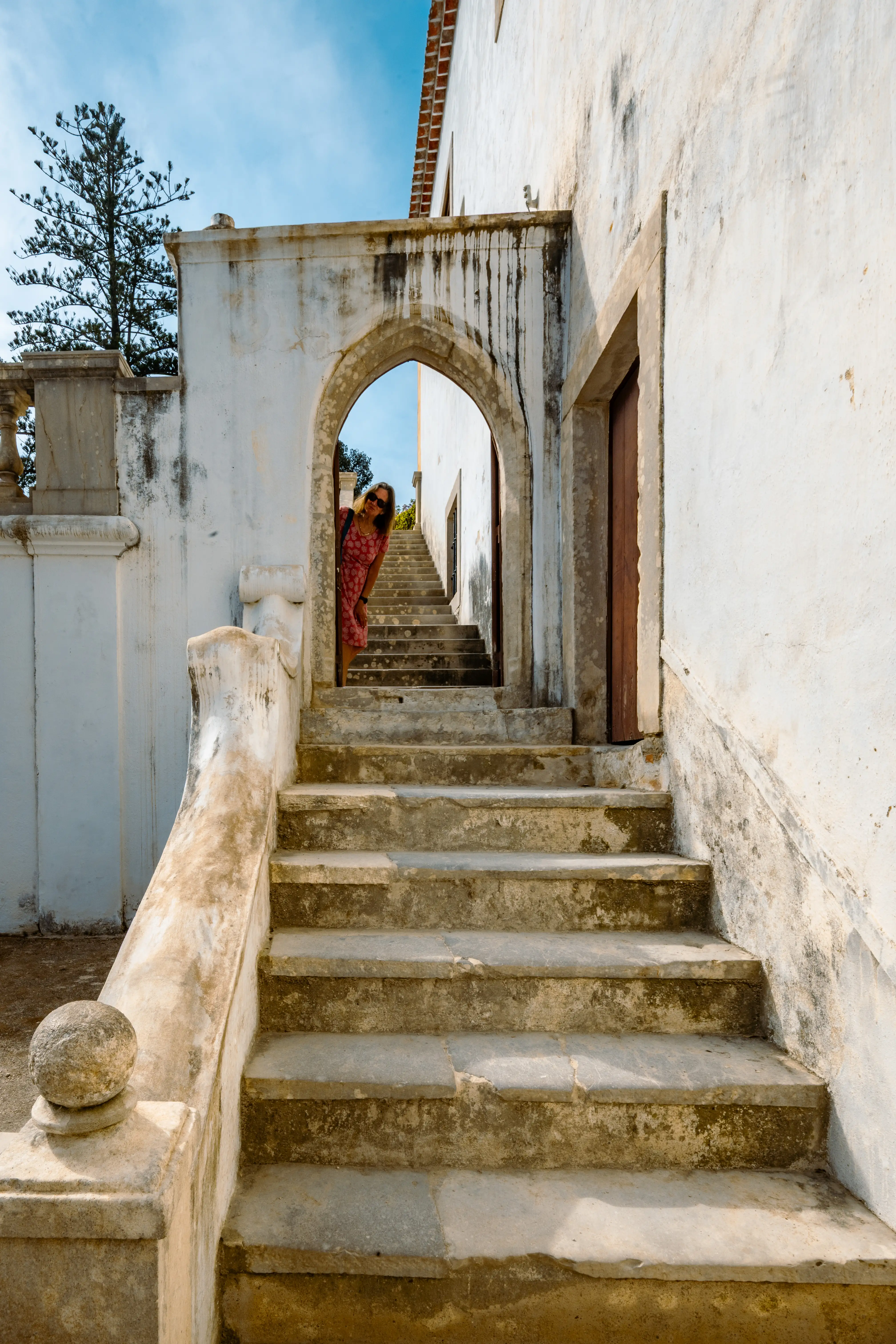 A woman wearing a red dress peeks around a white stone archway, overlooking an outdoor stone staircase.