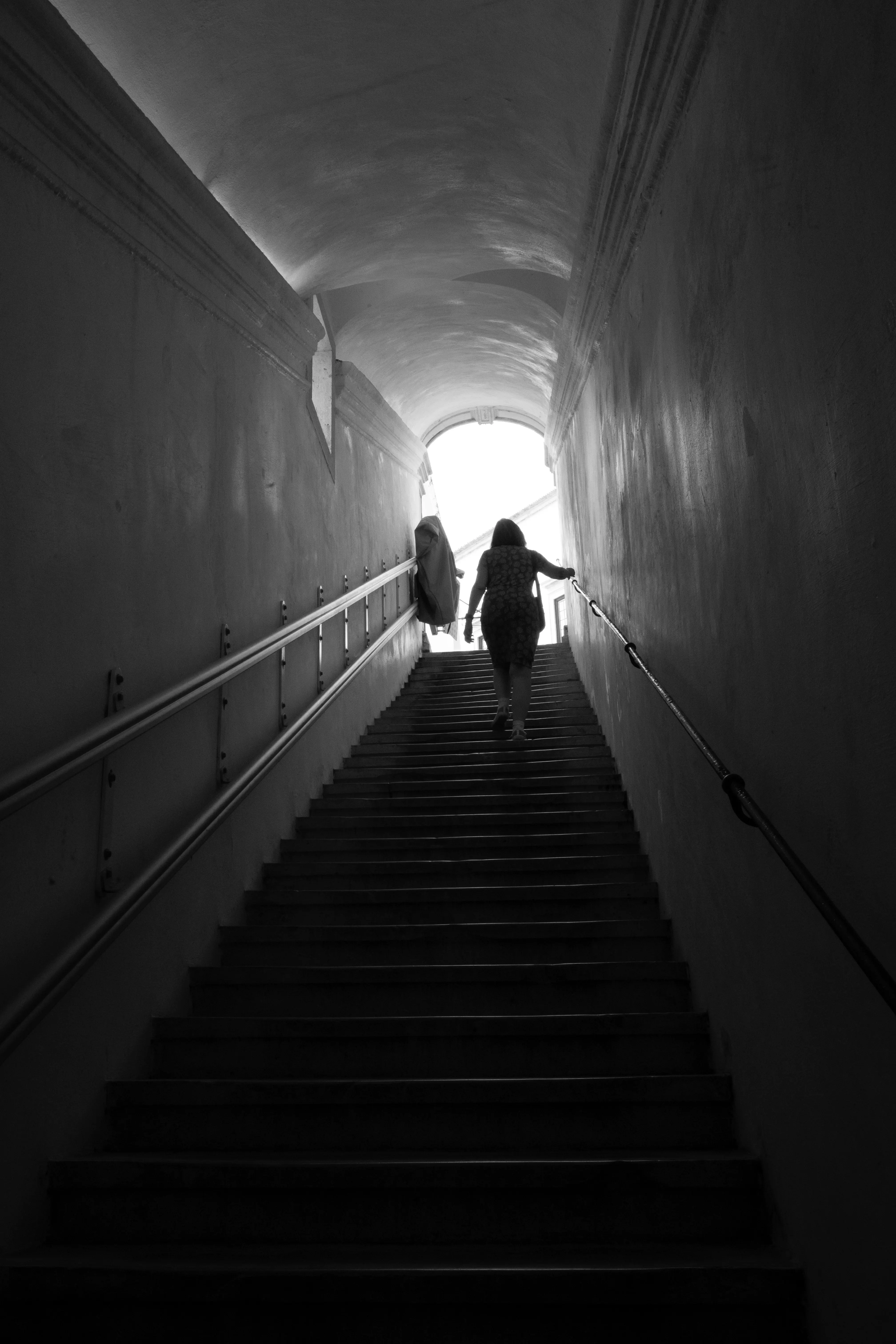 A silhouette of a woman in a red dress climbing a dark staircase towards a bright opening at the top, with metal handrails on either side.