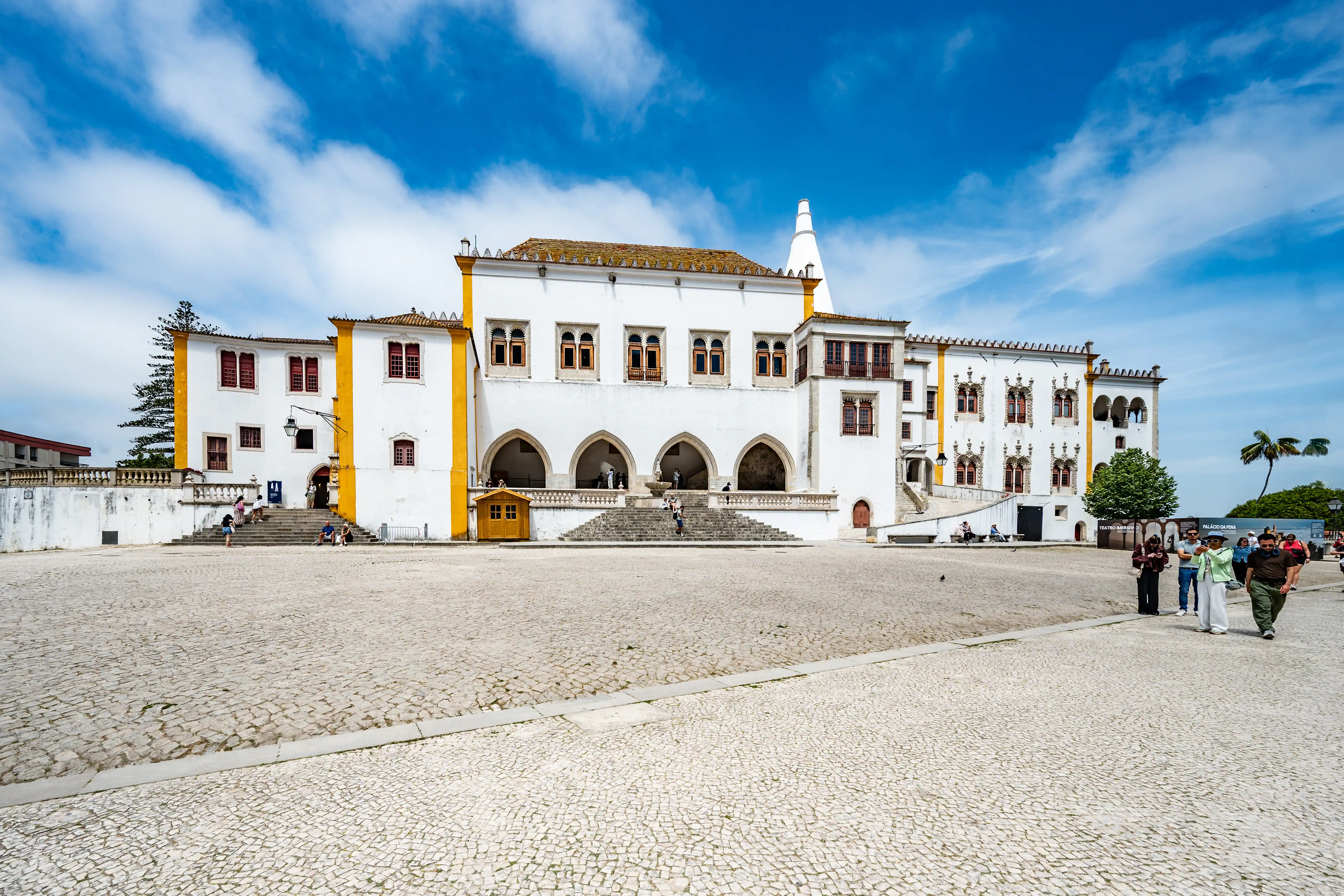 The white facade of Sintra National Palace with yellow trim and arched walkways. Several people are walking in the cobbled courtyard in front of the palace under a partly cloudy blue sky.