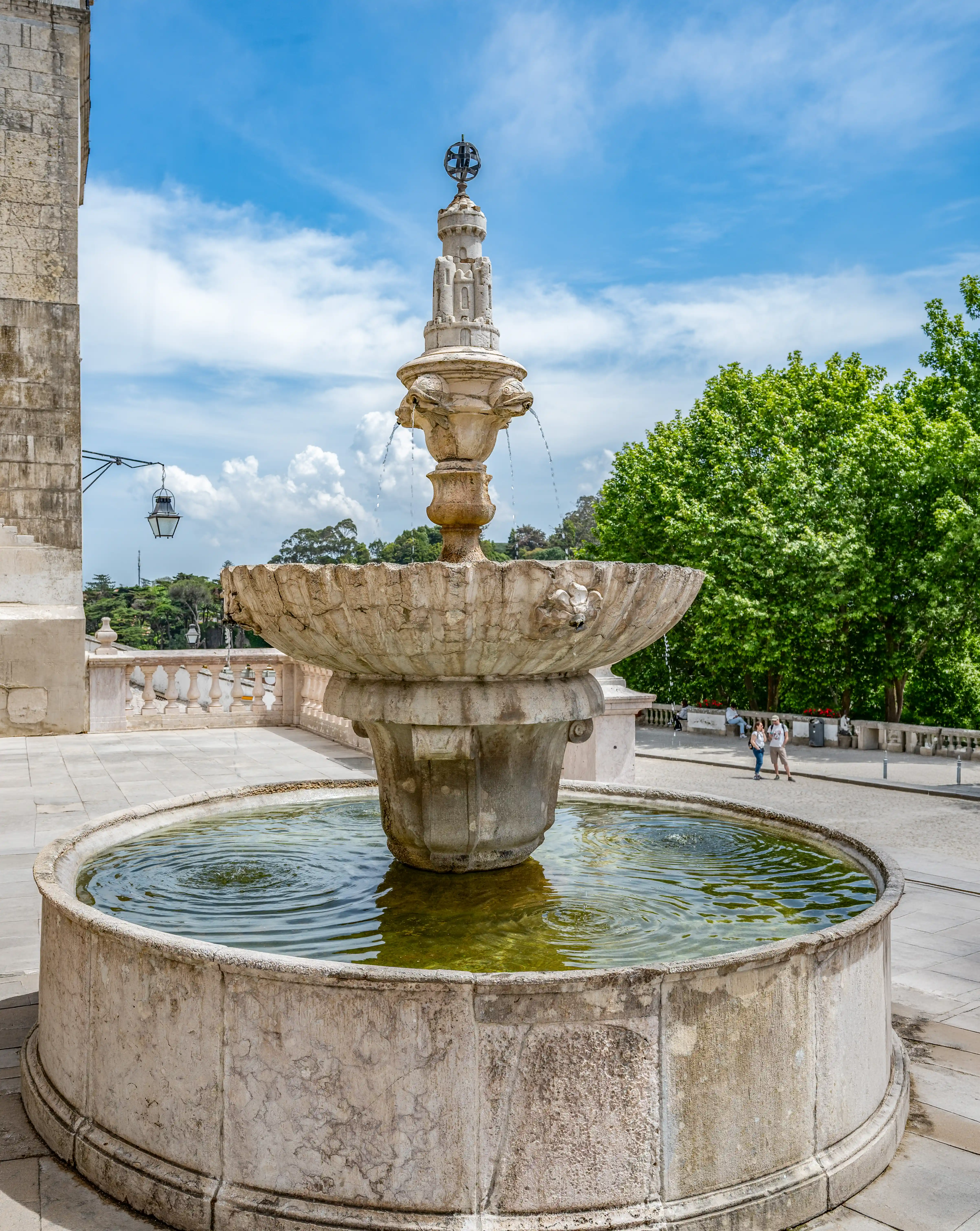 A detailed stone fountain with water flowing from decorative lion heads, set within a paved courtyard with a circular pool, a balustrade, and trees under a blue sky.