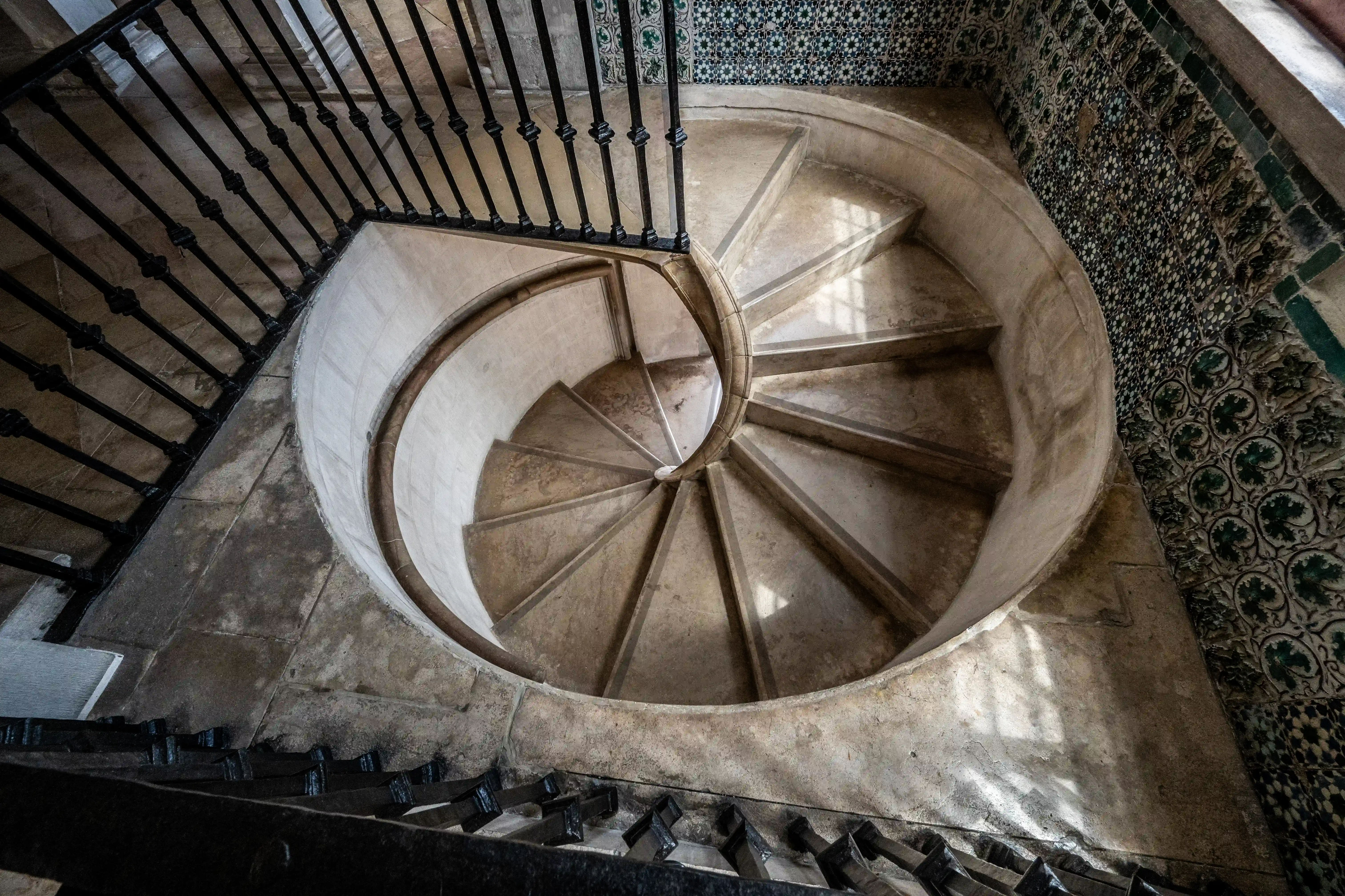 Overhead view of a stone spiral staircase with a dark metal balustrade and walls decorated with traditional blue and green patterned azulejo tiles. A small, barred window is visible on the landing.