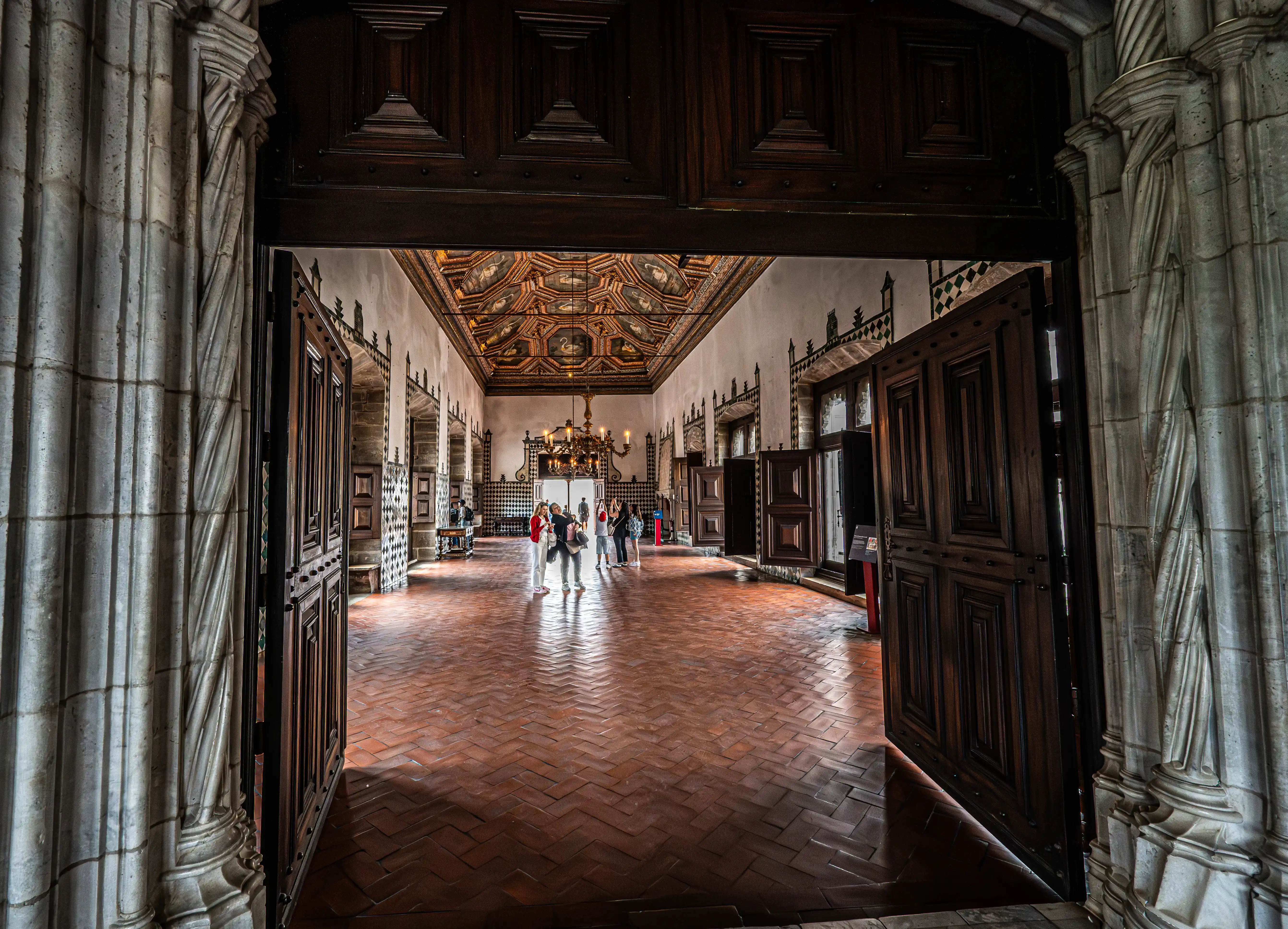 A wide view of a long, historic hallway seen through a grand arched entrance. Ornate tiled walls, a brick floor, and a detailed ceiling are visible, with a chandelier in the distance. Several people are walking in the hallway.