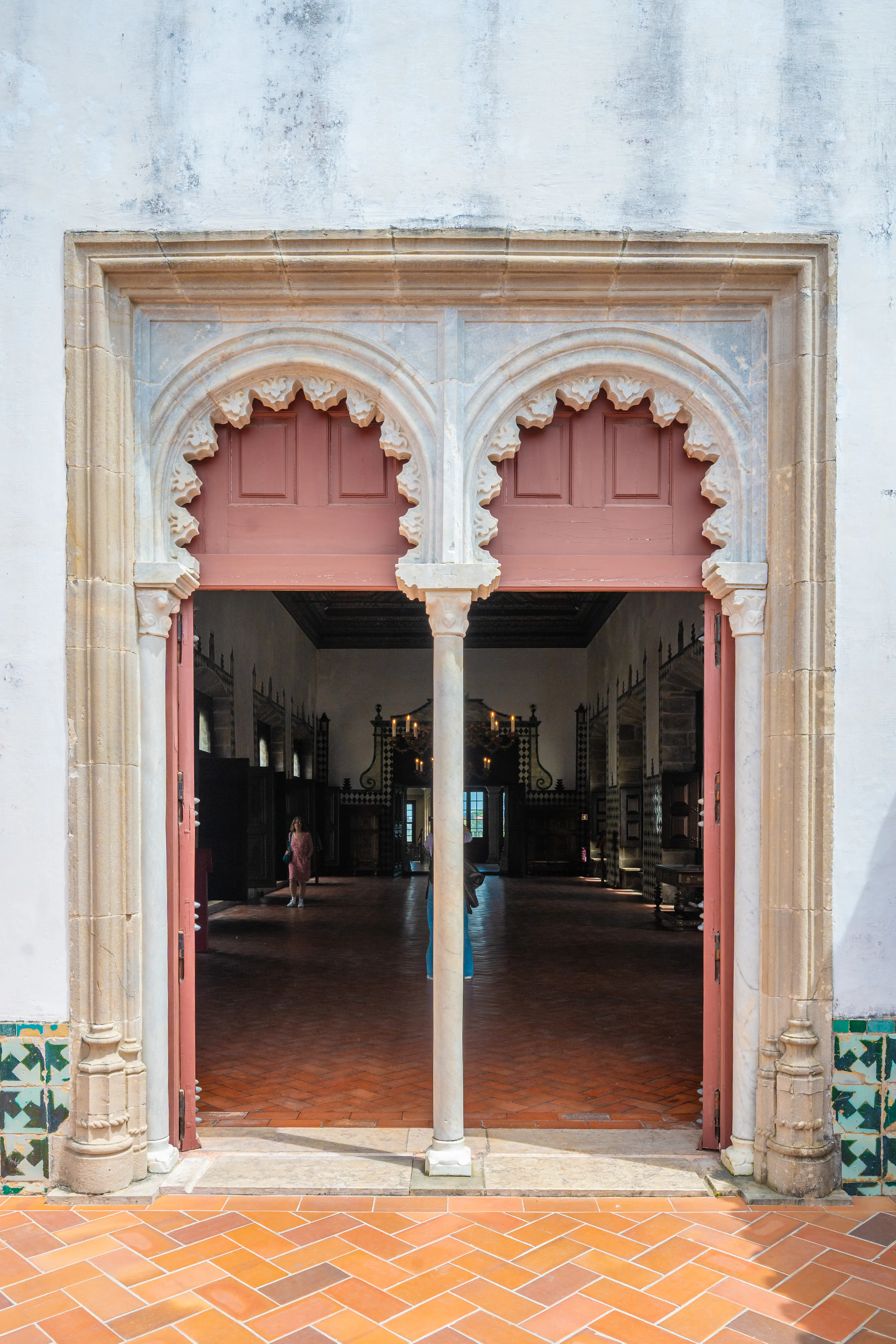 A detailed view of a stone carved double archway with two dark wooden doors partially open, leading into a shadowy interior. The foreground shows a patterned terracotta tile floor and decorative tiled walls flanking the entrance.