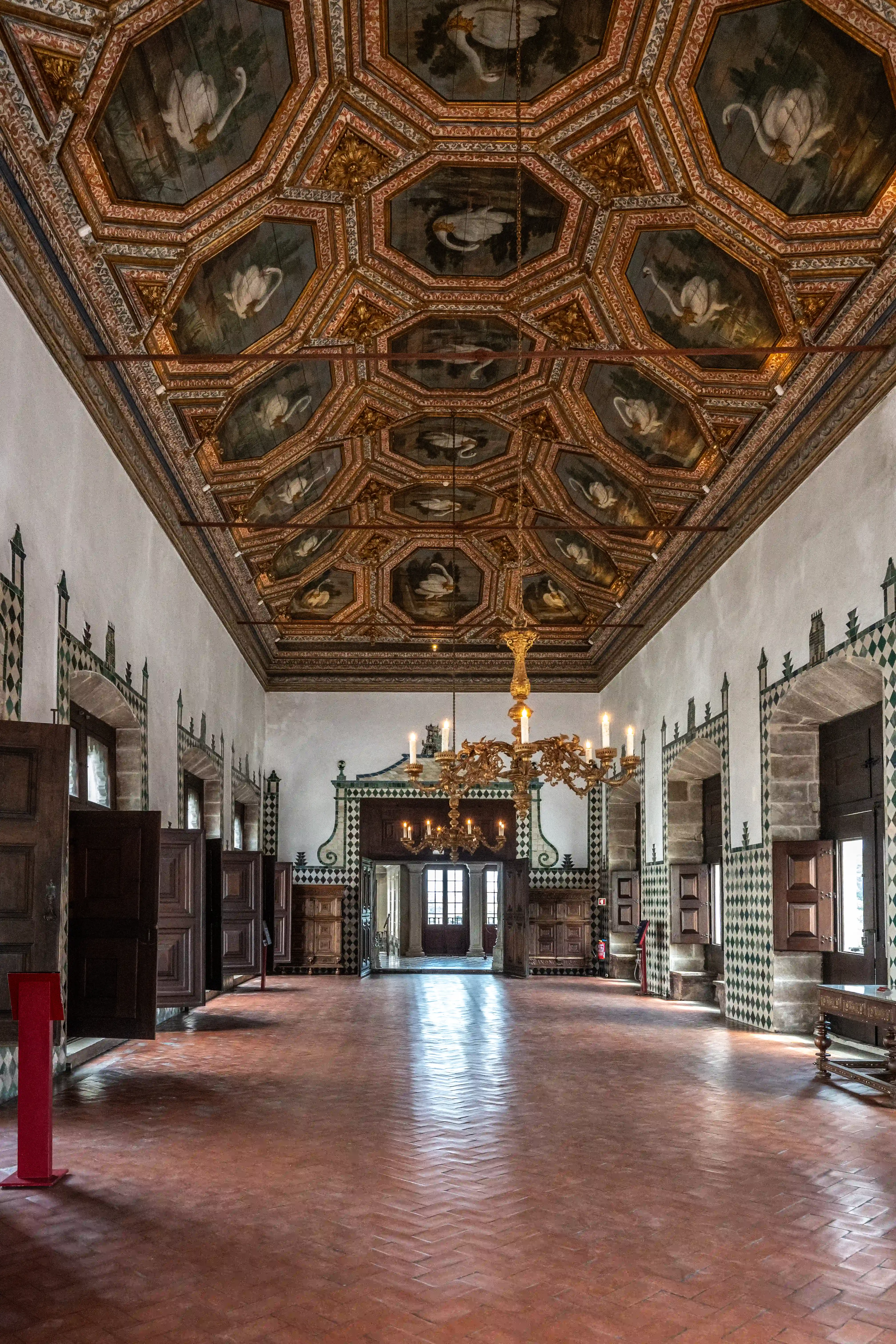  A grand hall with a detailed painted ceiling showing swans, ornate walls, a large chandelier, and a herringbone tiled floor.